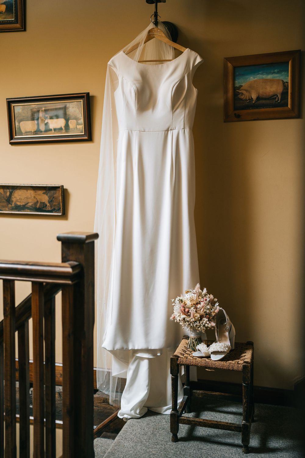A white wedding dress hangs elegantly against a beige wall with framed artwork and wooden railing details.