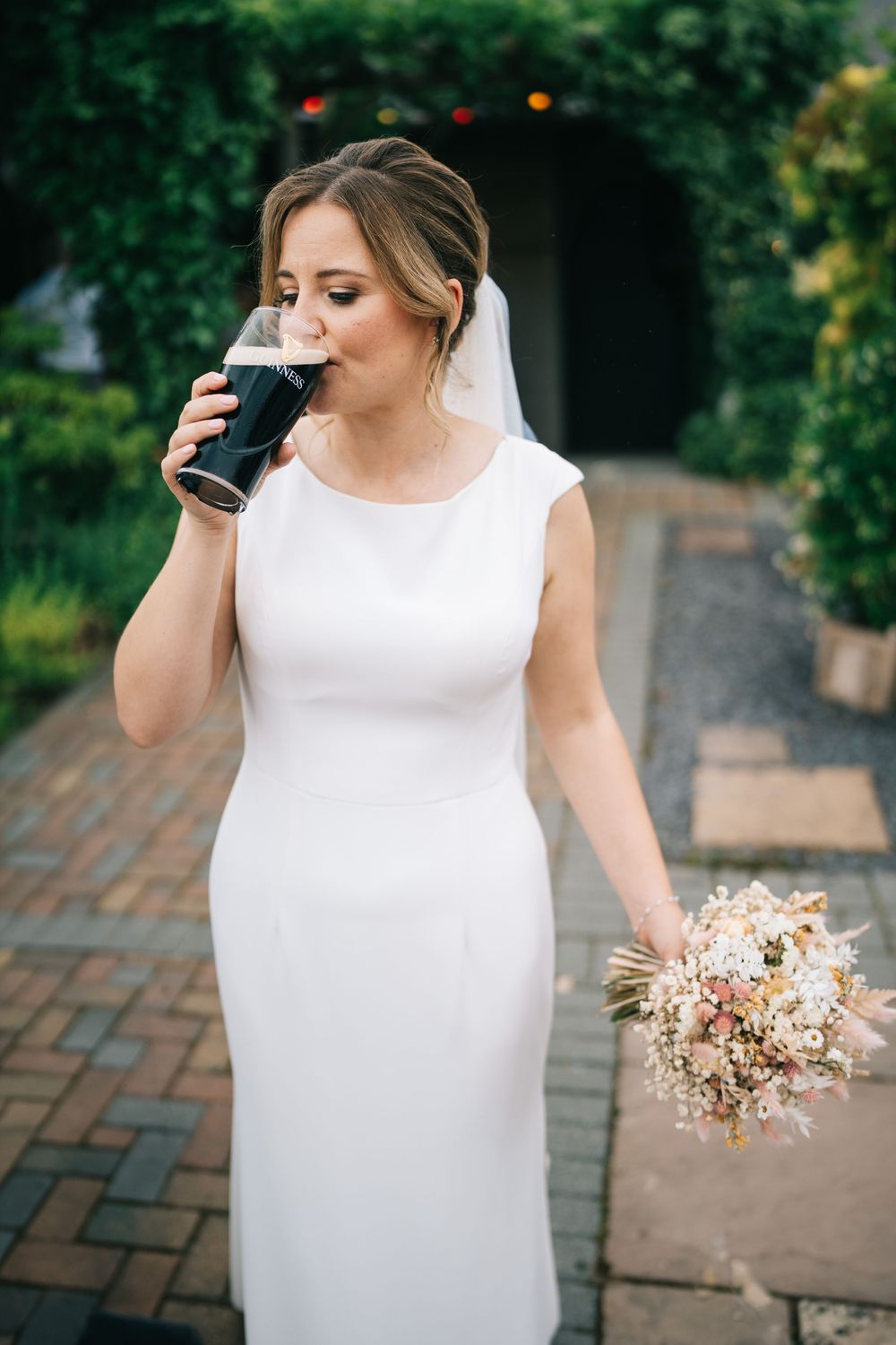 A bride in a sleeveless white dress holds a delicate pink and white bouquet while walking on brick pavement, Bromwich Park Farm, Shropshire