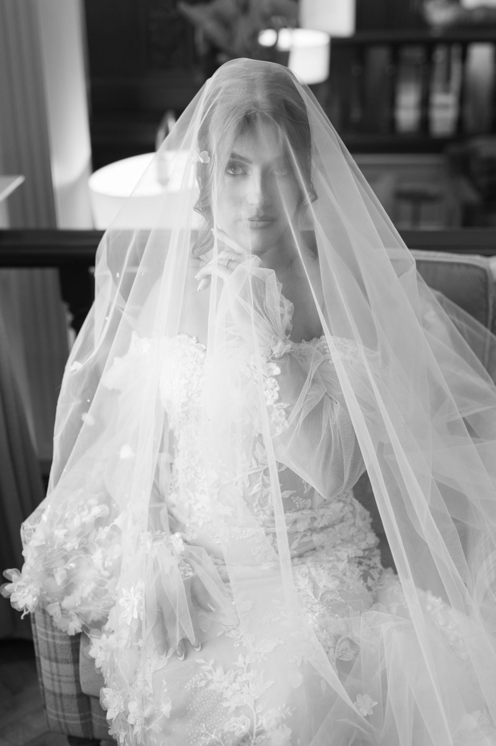 Black and white portrait of a bride wearing a floral lace gown and flowing veil over her face.