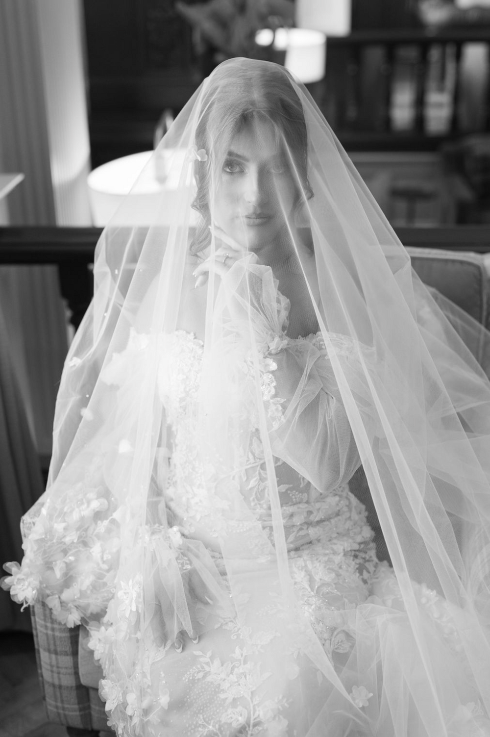 Black and white portrait of a bride wearing a floral lace gown and flowing veil over her face.