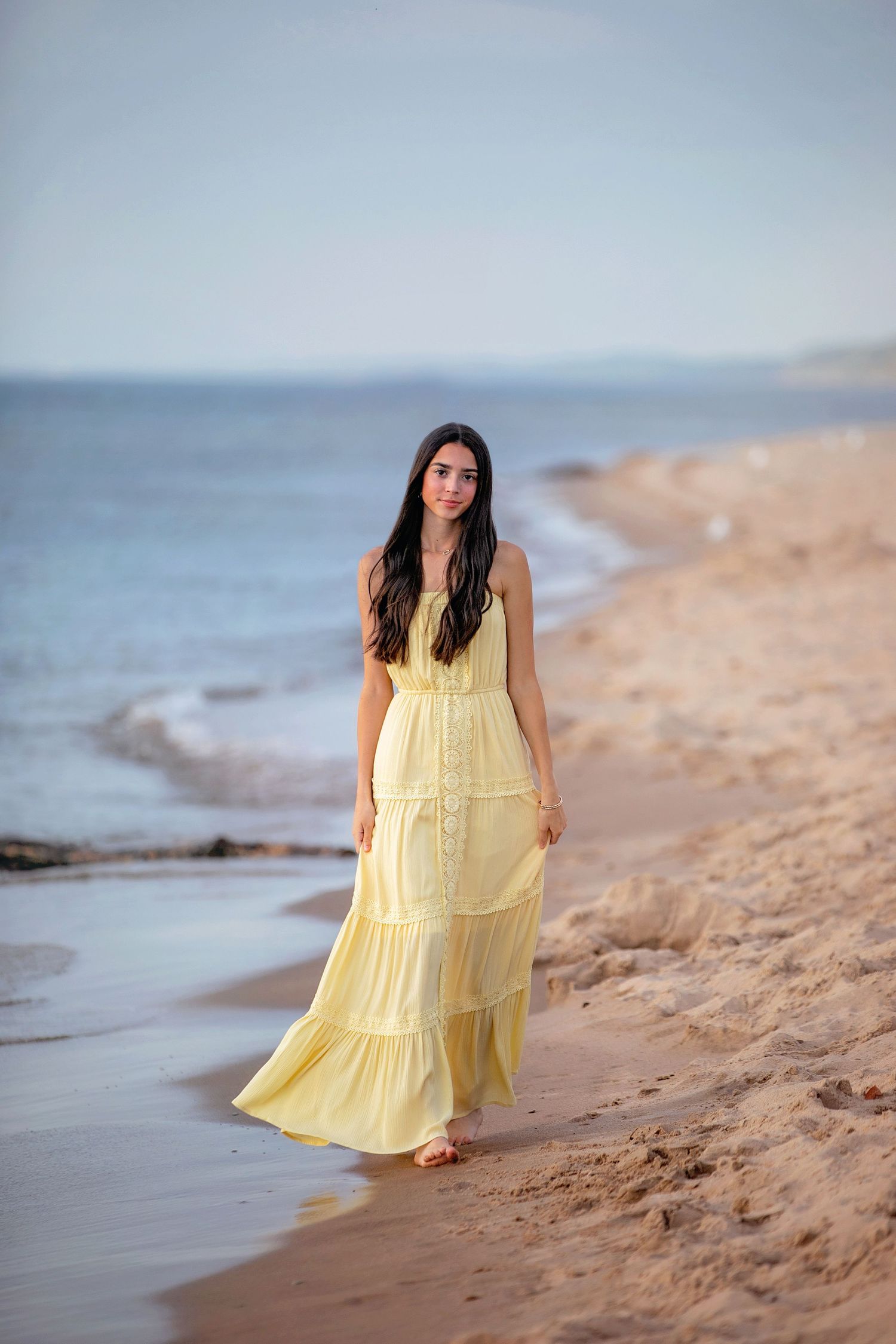 A person in a flowing yellow maxi dress walks along a peaceful beach at sunset with ocean waves in the background.