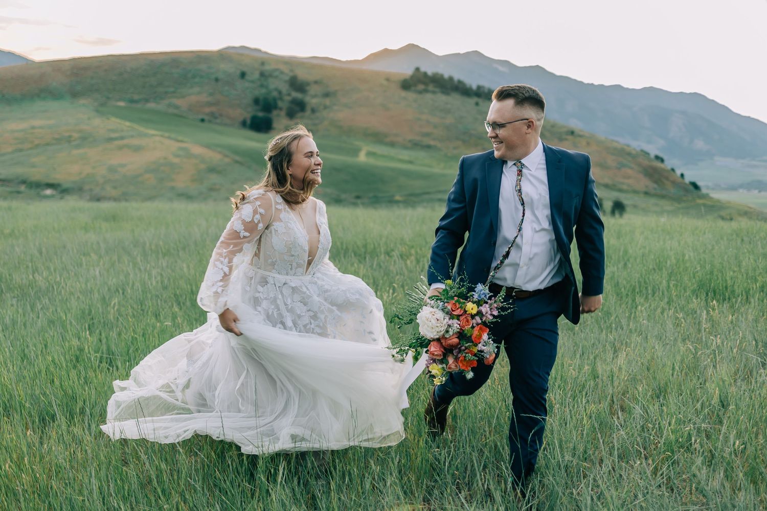 Bride and groom walking through mountain meadow with colorful bouquet.