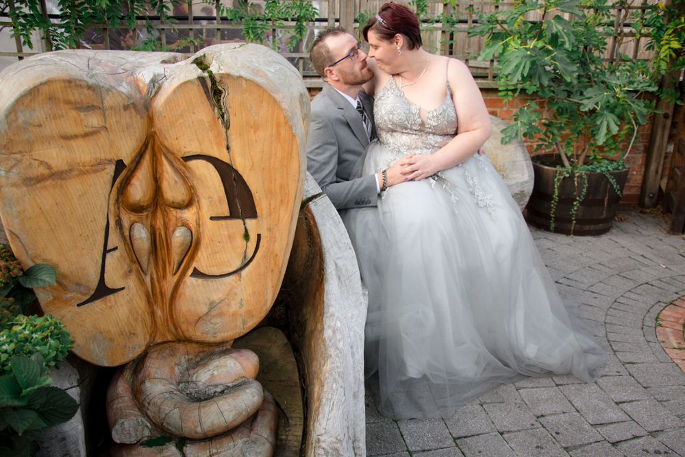 A couple in wedding attire share an intimate moment next to a decorative stone wall sculpture in a garden setting.