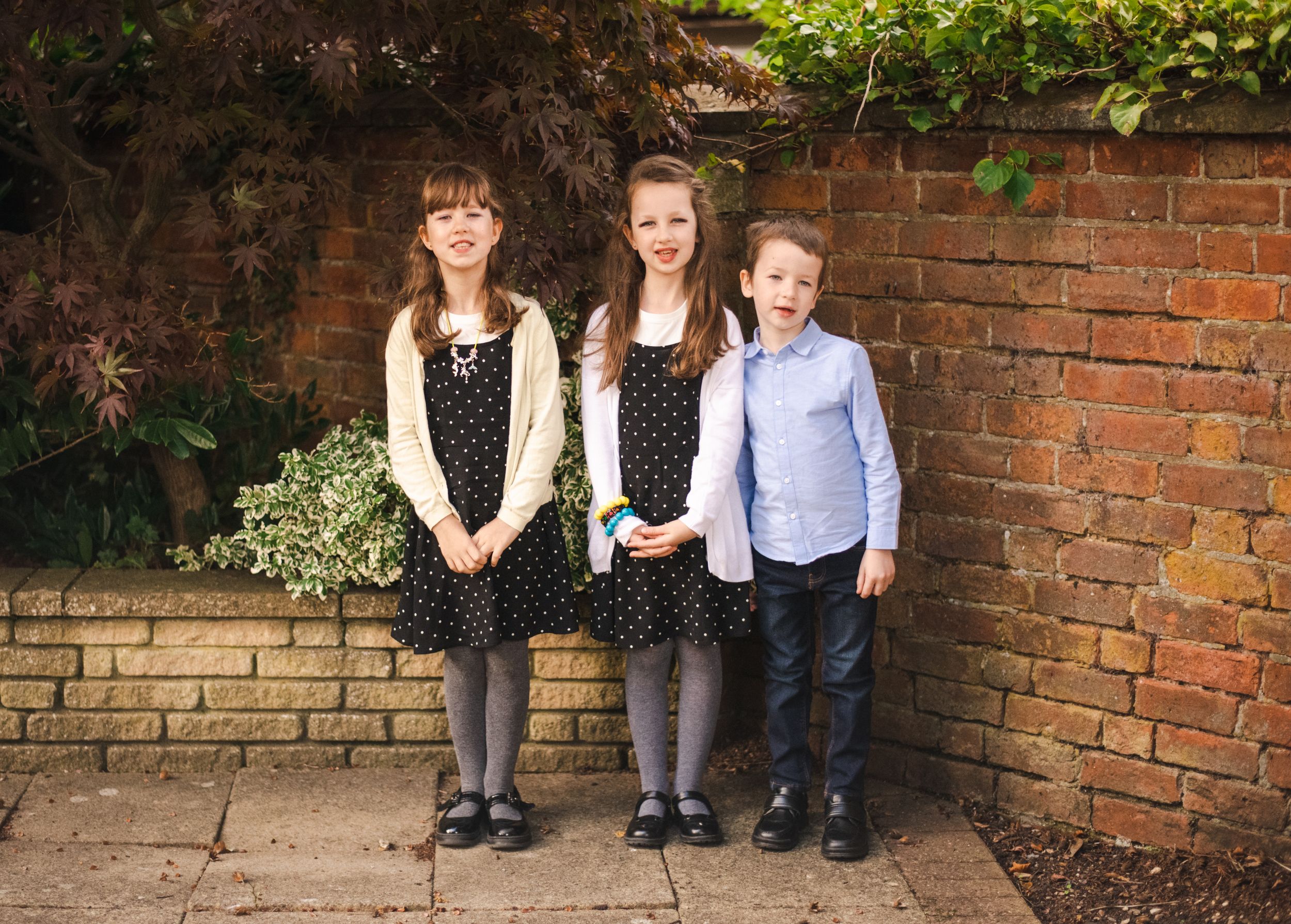 Children in formal attire pose against a brick wall outdoors.