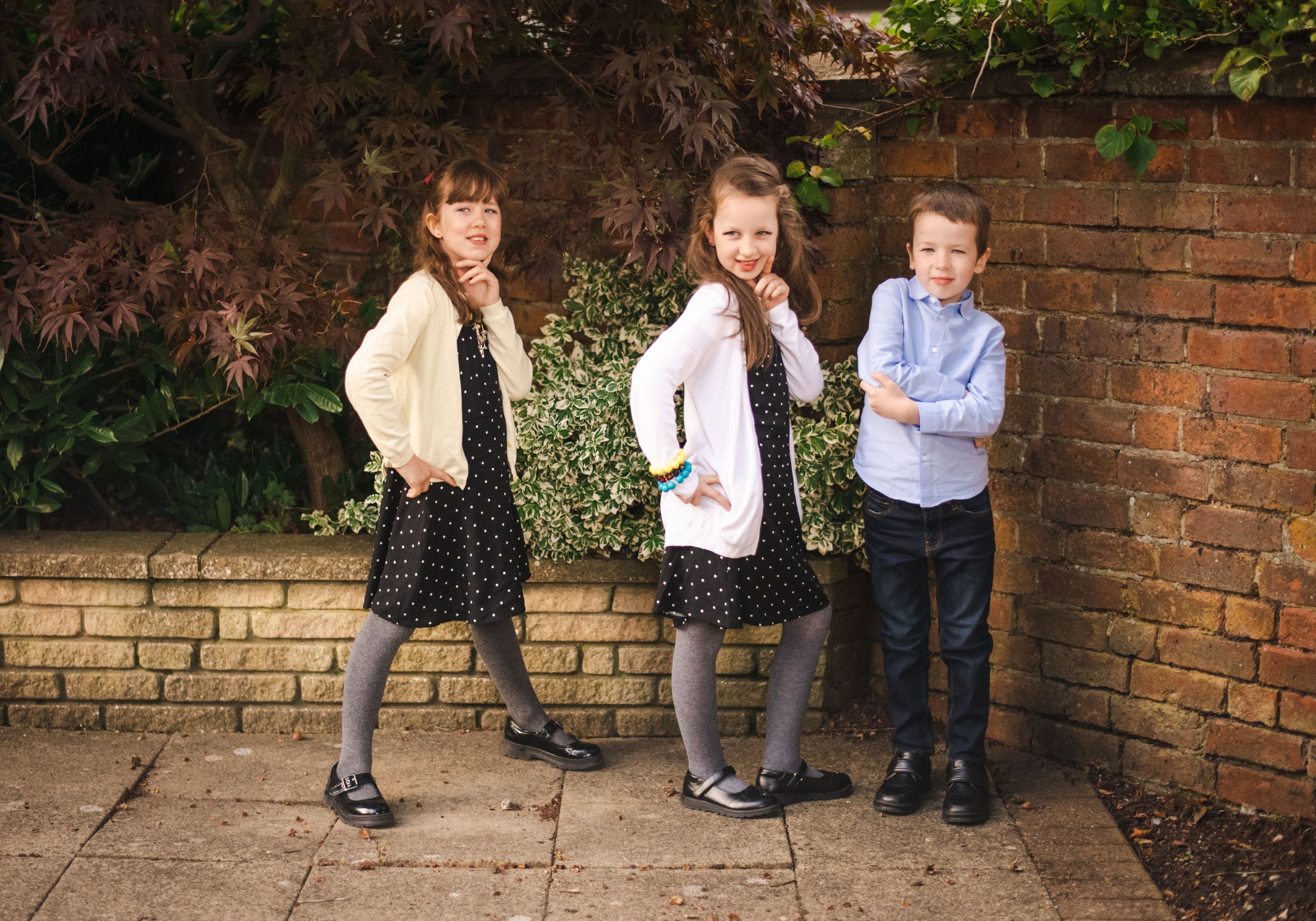 Three children pose casually against a brick wall in school uniforms.