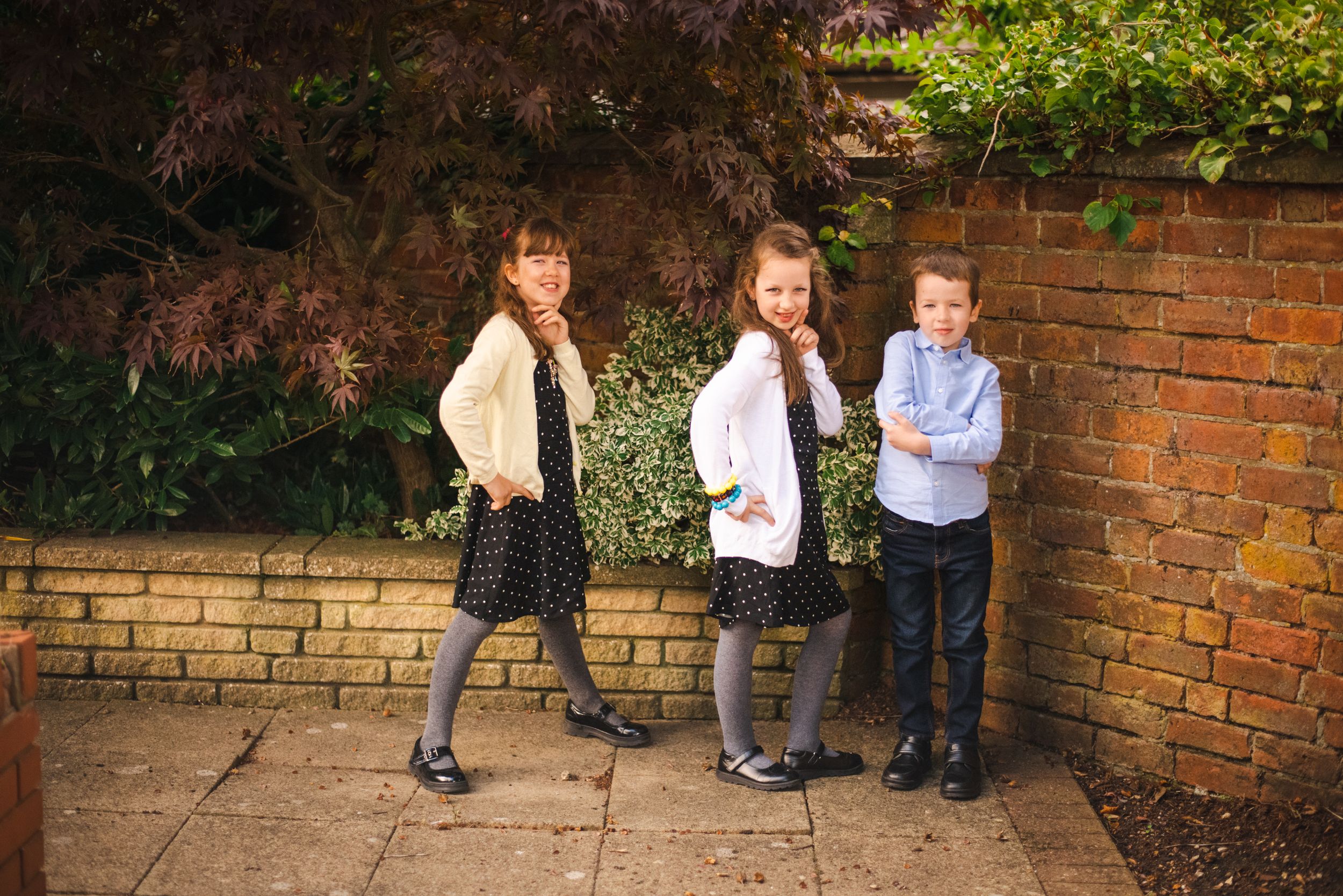 Three young students lean against a garden brick wall outdoors.