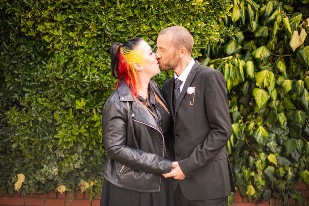 A couple shares an intimate moment in front of a lush green hedge.