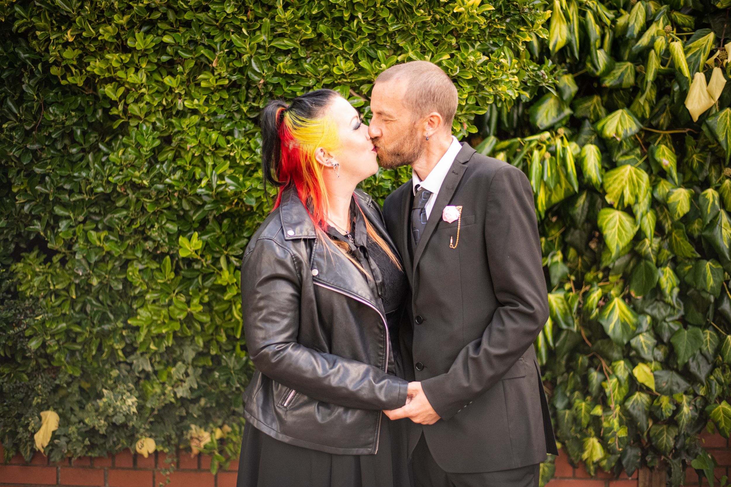A couple shares an intimate moment in front of a lush green hedge.