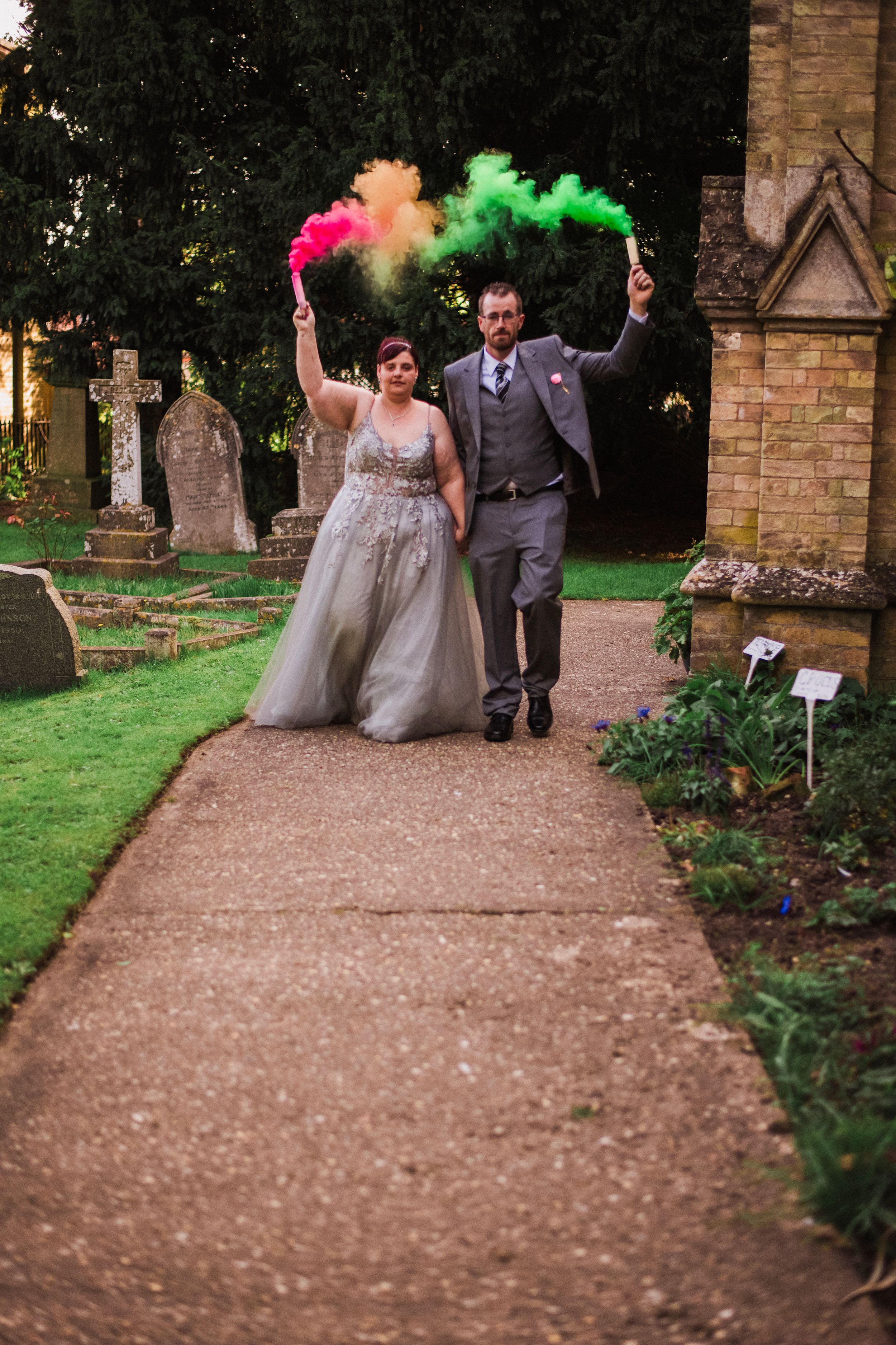Newlyweds celebrate with colorful smoke bombs on a church pathway.