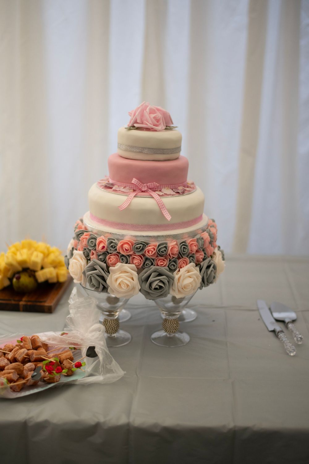 Elegant three-tier wedding cake decorated with pink and gray roses on a crystal stand.