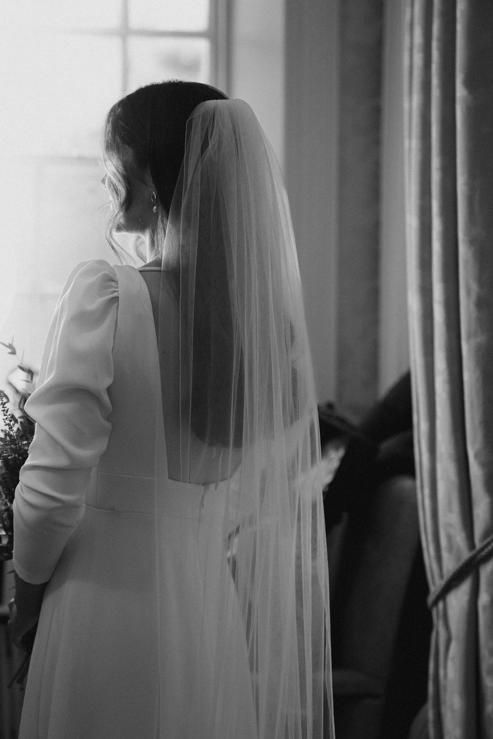 Back view of bride in long white dress and flowing veil standing by window in soft light.
