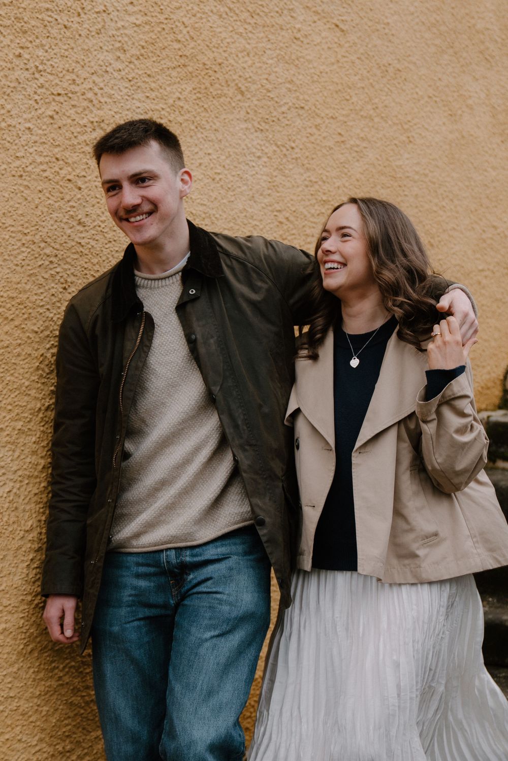 A smiling young couple in casual jackets laughing together against a yellow textured wall outdoors.