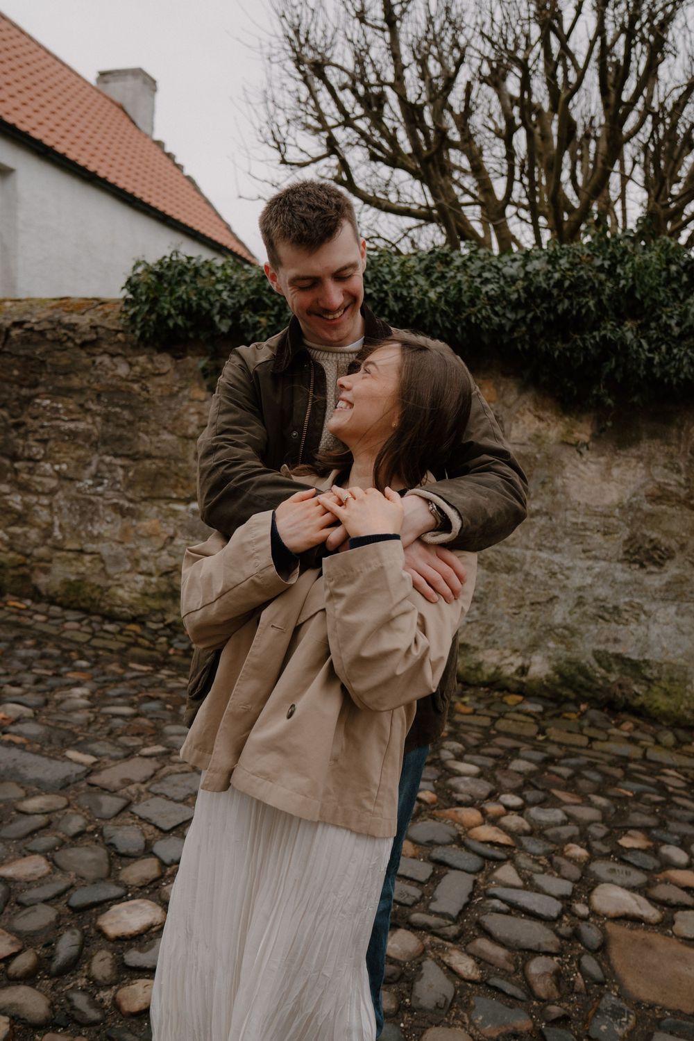 A happy couple embracing outdoors near a stone wall, smiling together in casual autumn attire.