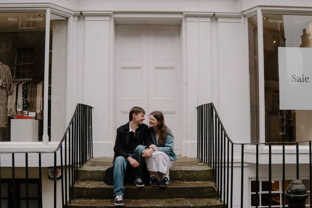 Couple sitting on steps outside a white storefront, smiling at each other in a candid romantic moment.