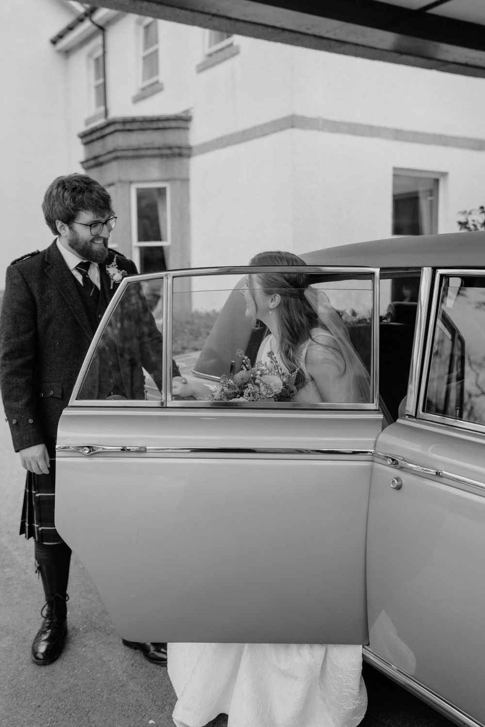 Black and white photo of a groom in a kilt holding the car door open for a bride in a wedding dress.
