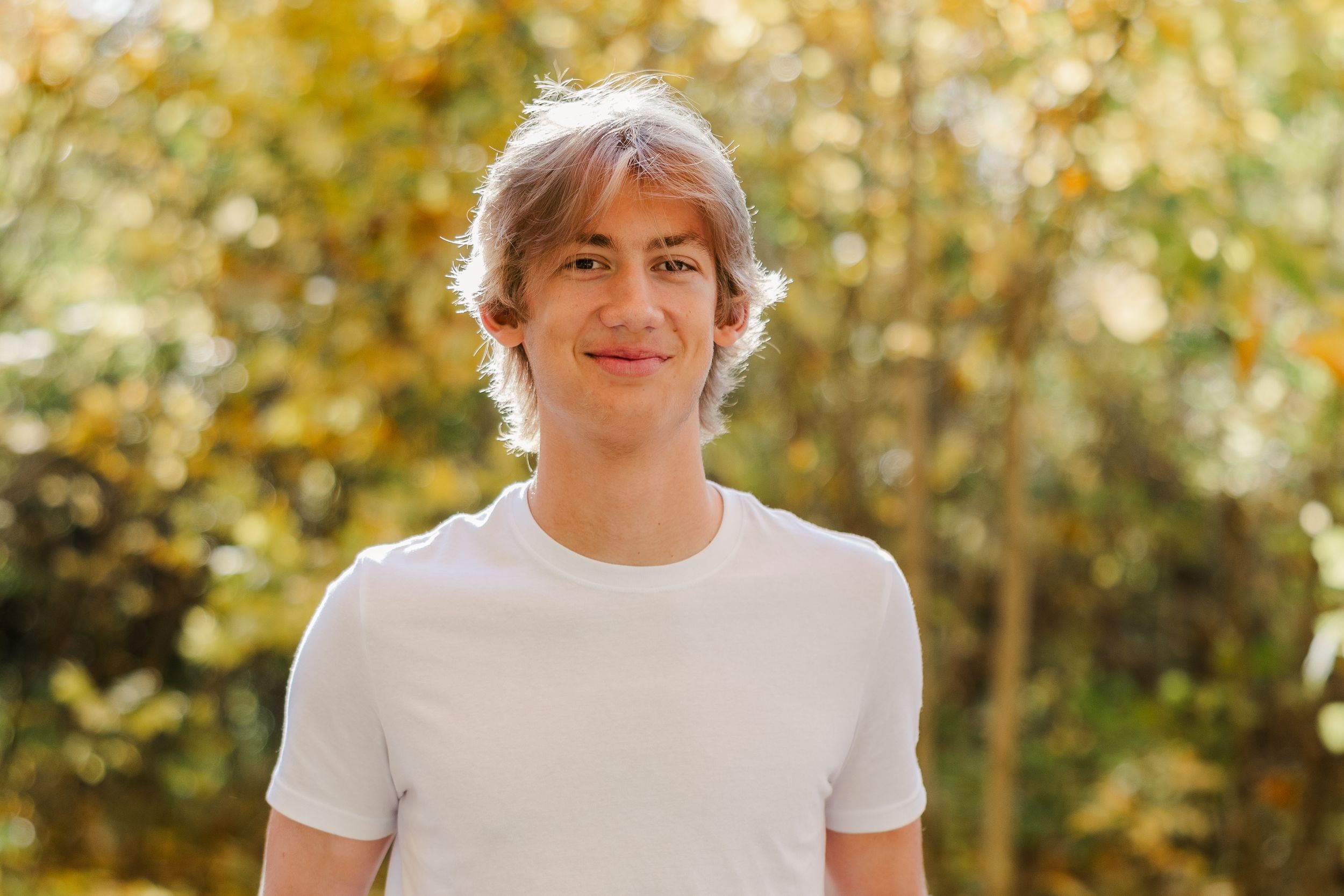 High school senior standing and smiling on a pathway surrounded by vibrant fall foliage.
