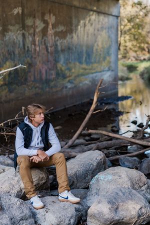High school boy sitting on large boulders in the middle of the river under a bridge.