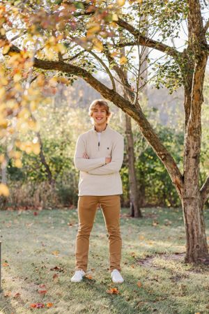High school senior standing with his arms crossed under a tree with vibrant yellow leaves.