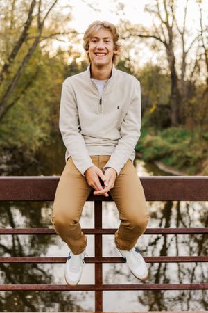 High school senior sitting on a wooden and metal bridge surrounded by fall colors.
