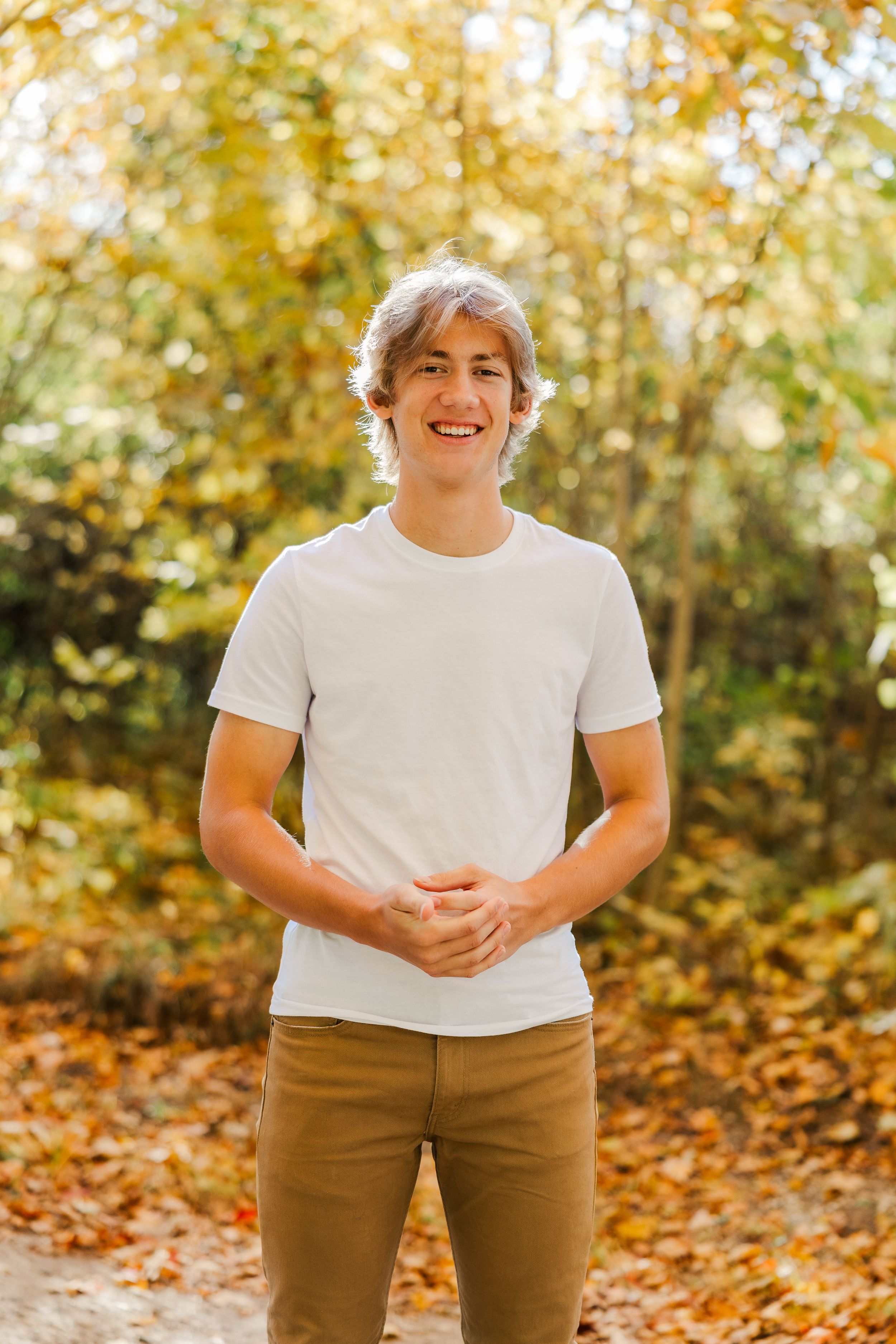 High school senior standing and smiling on a pathway surrounded by vibrant fall foliage.