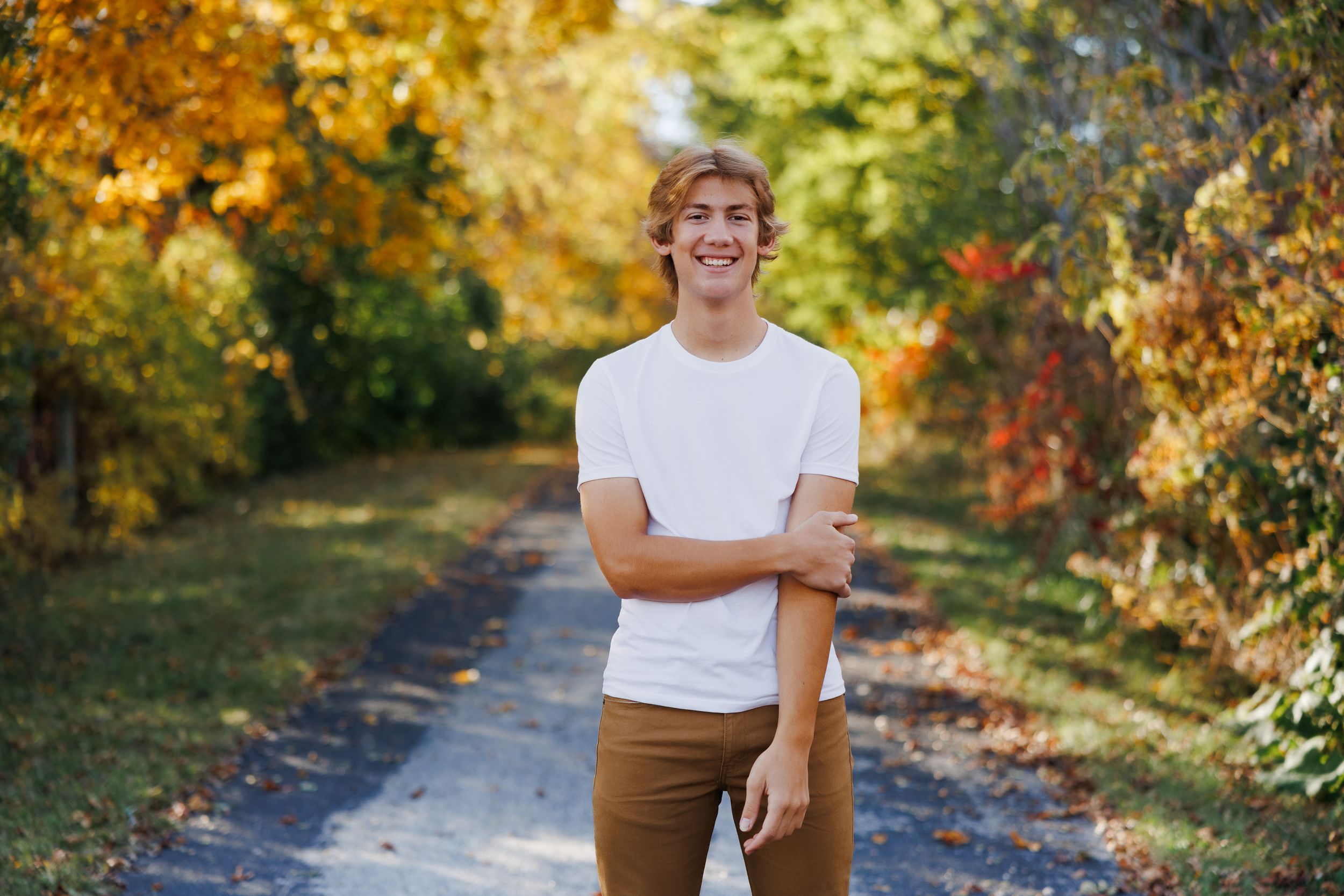 High school senior standing and smiling on a pathway surrounded by vibrant fall foliage.