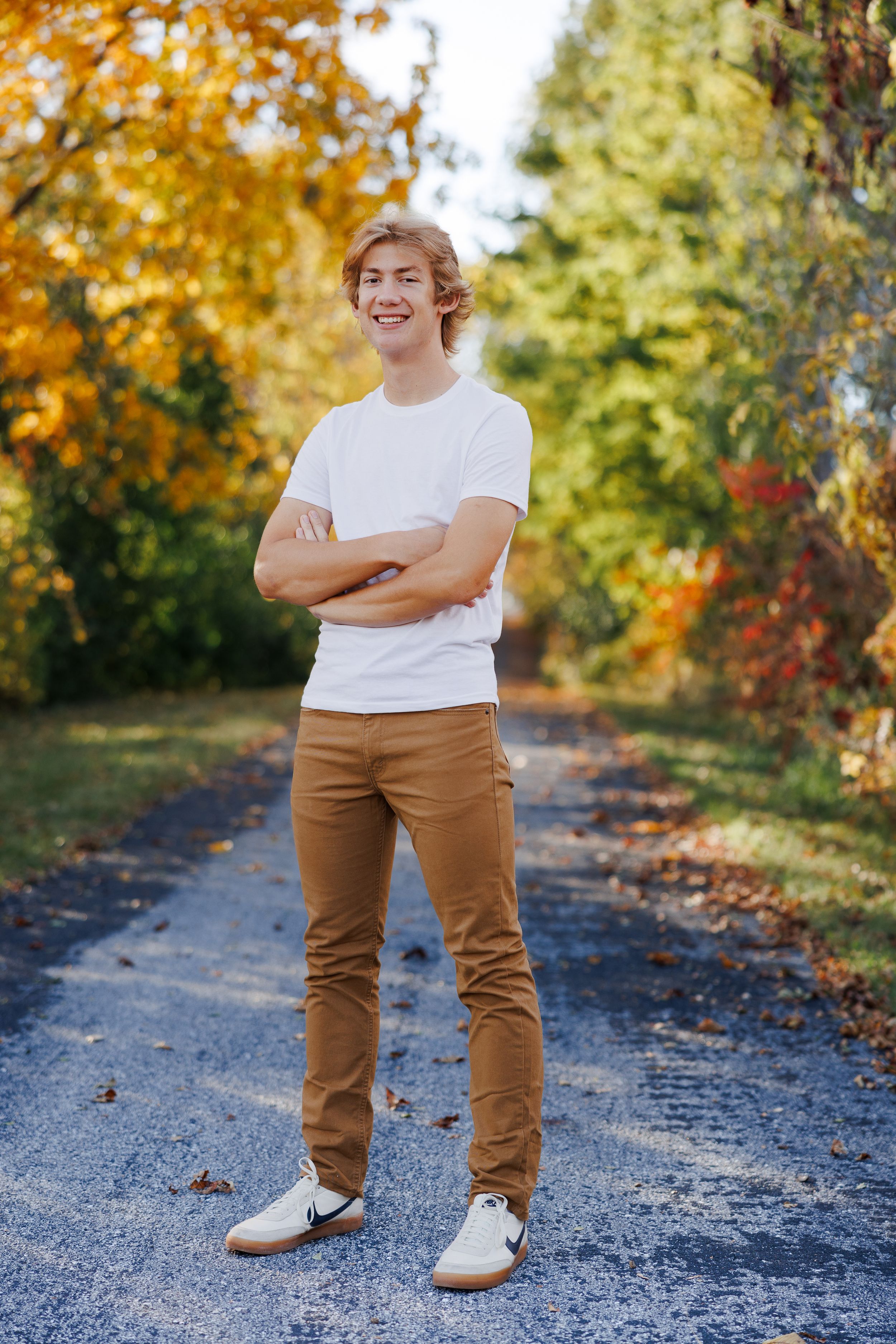 High school senior standing and smiling on a pathway surrounded by vibrant fall foliage.