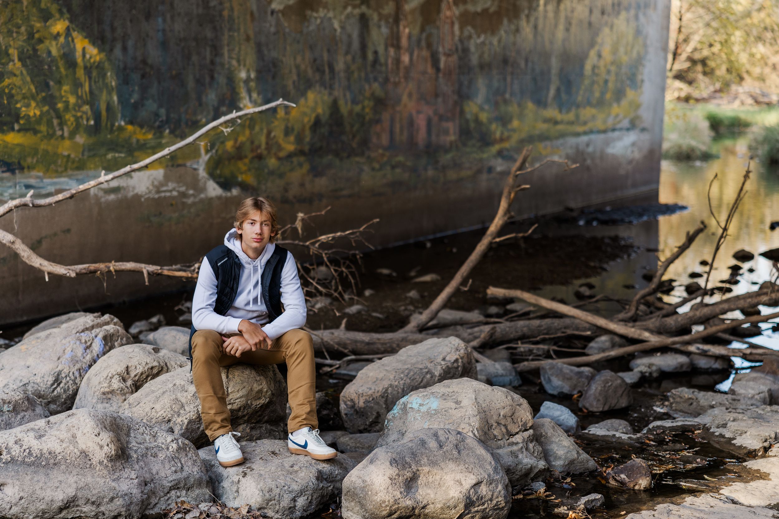 High school boy sitting on large boulders in the middle of the river under a bridge.