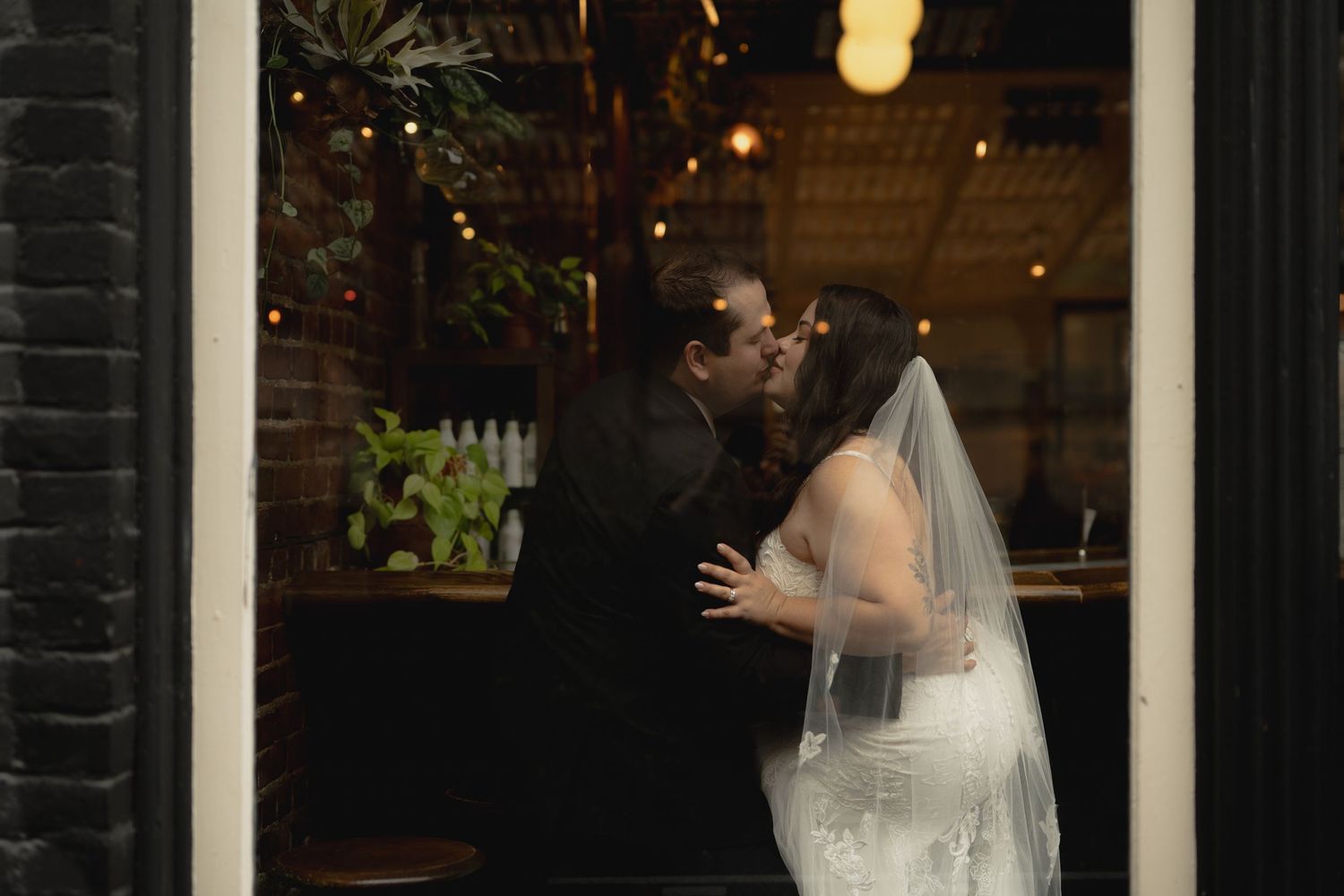 A tender wedding moment captured through a window with string lights glowing in the background.