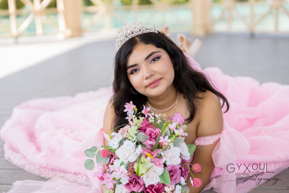A close-up portrait featuring a pink quinceañera gown and floral bouquet on an outdoor deck.