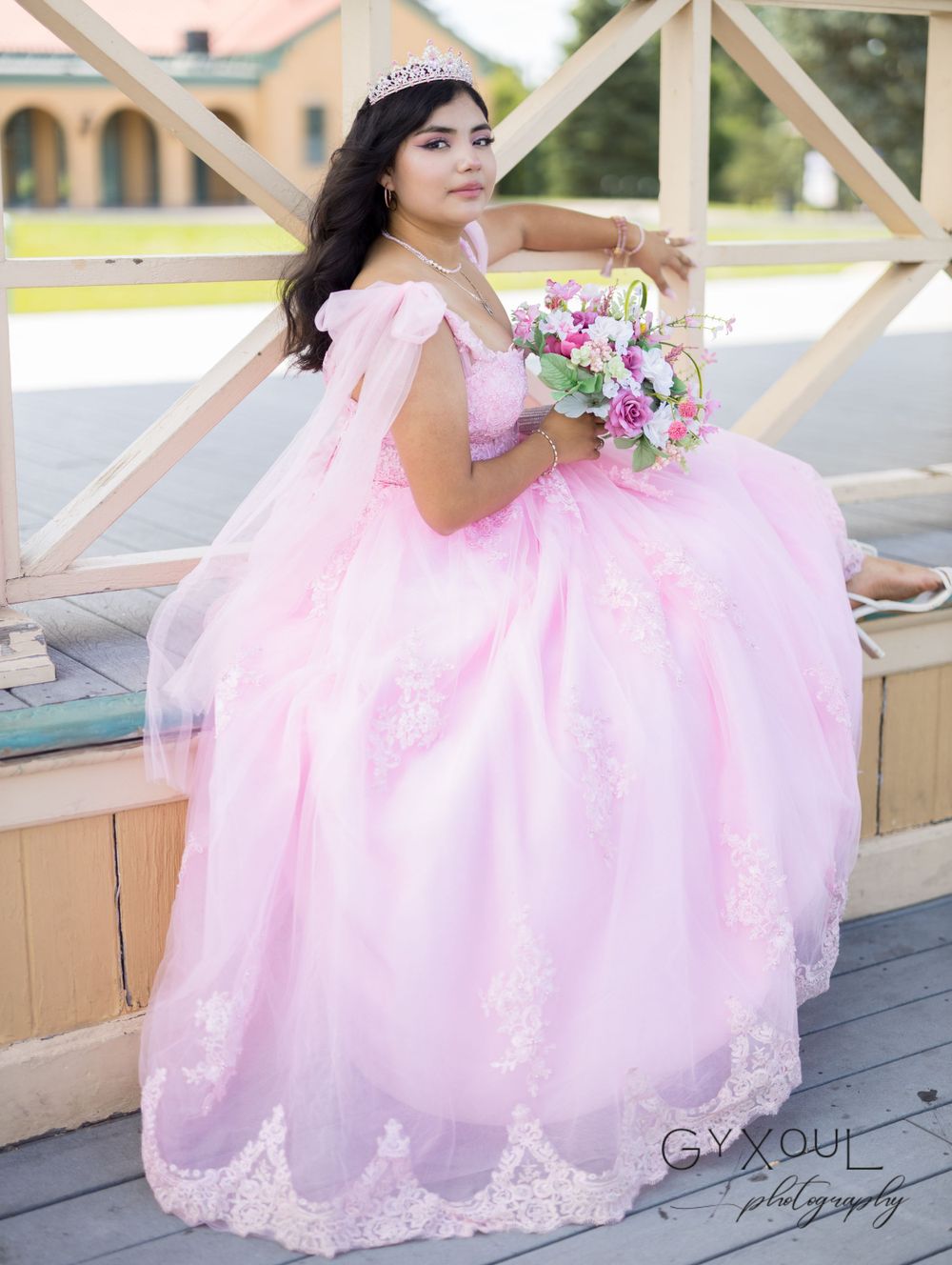 A portrait of someone in a pink lace ball gown sitting on a wooden deck railing.