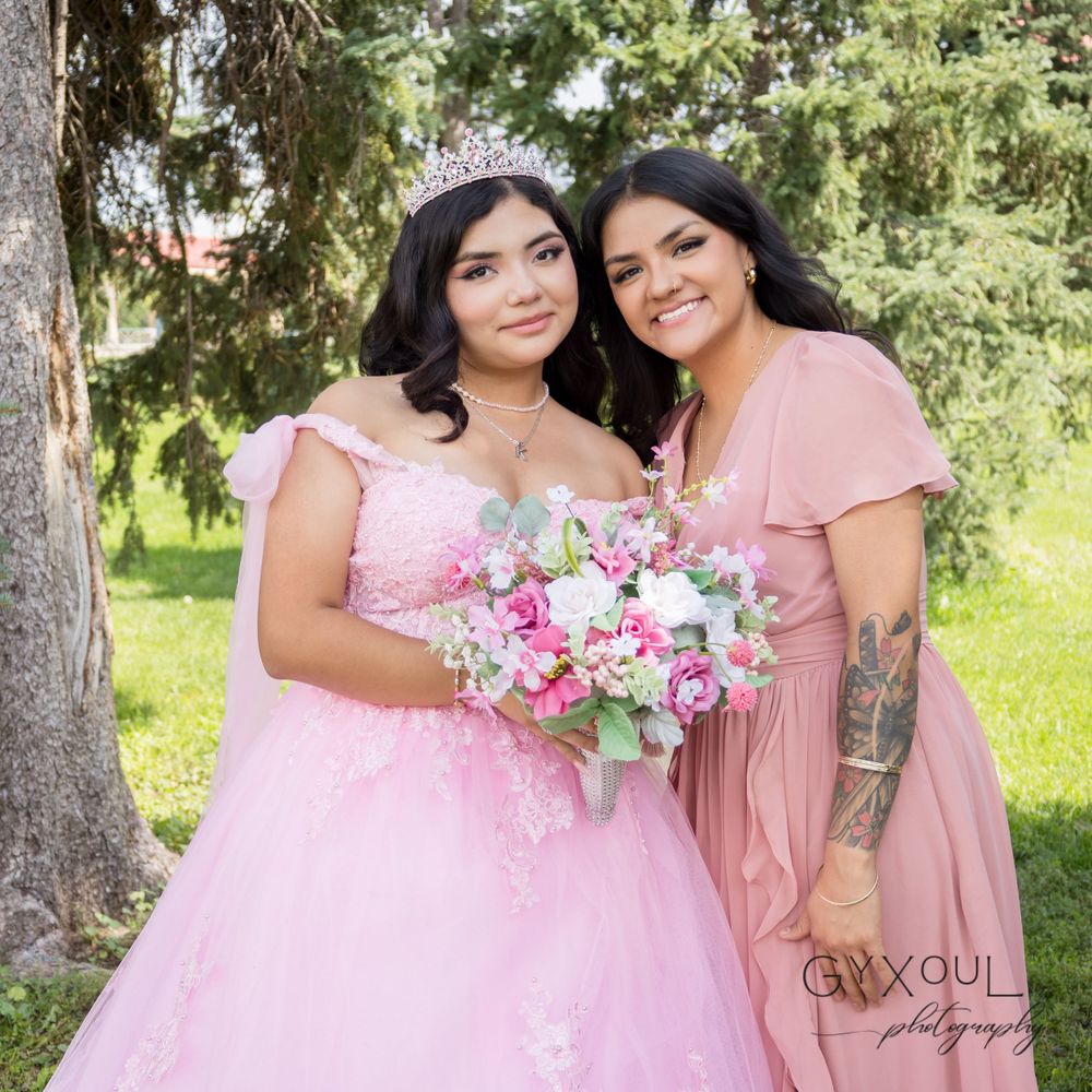 Two people in pink dresses pose with a floral bouquet during a quinceañera celebration outdoors.