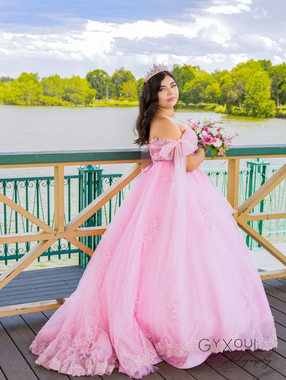 A pink ball gown photoshoot on a lakeside deck with lush greenery in the background.