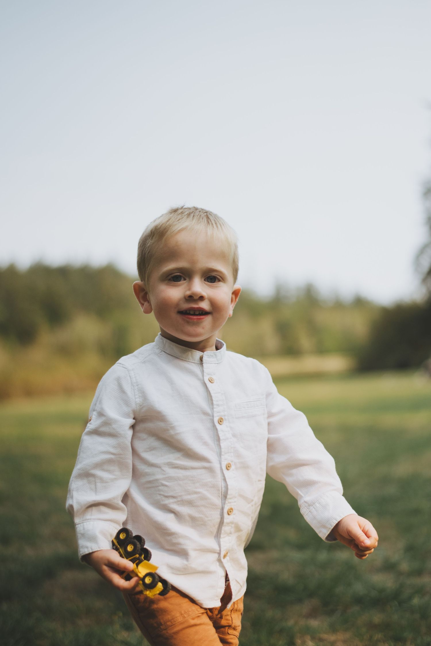 Young boy smiling and holding a toy truck while playing outdoors in a grassy field.