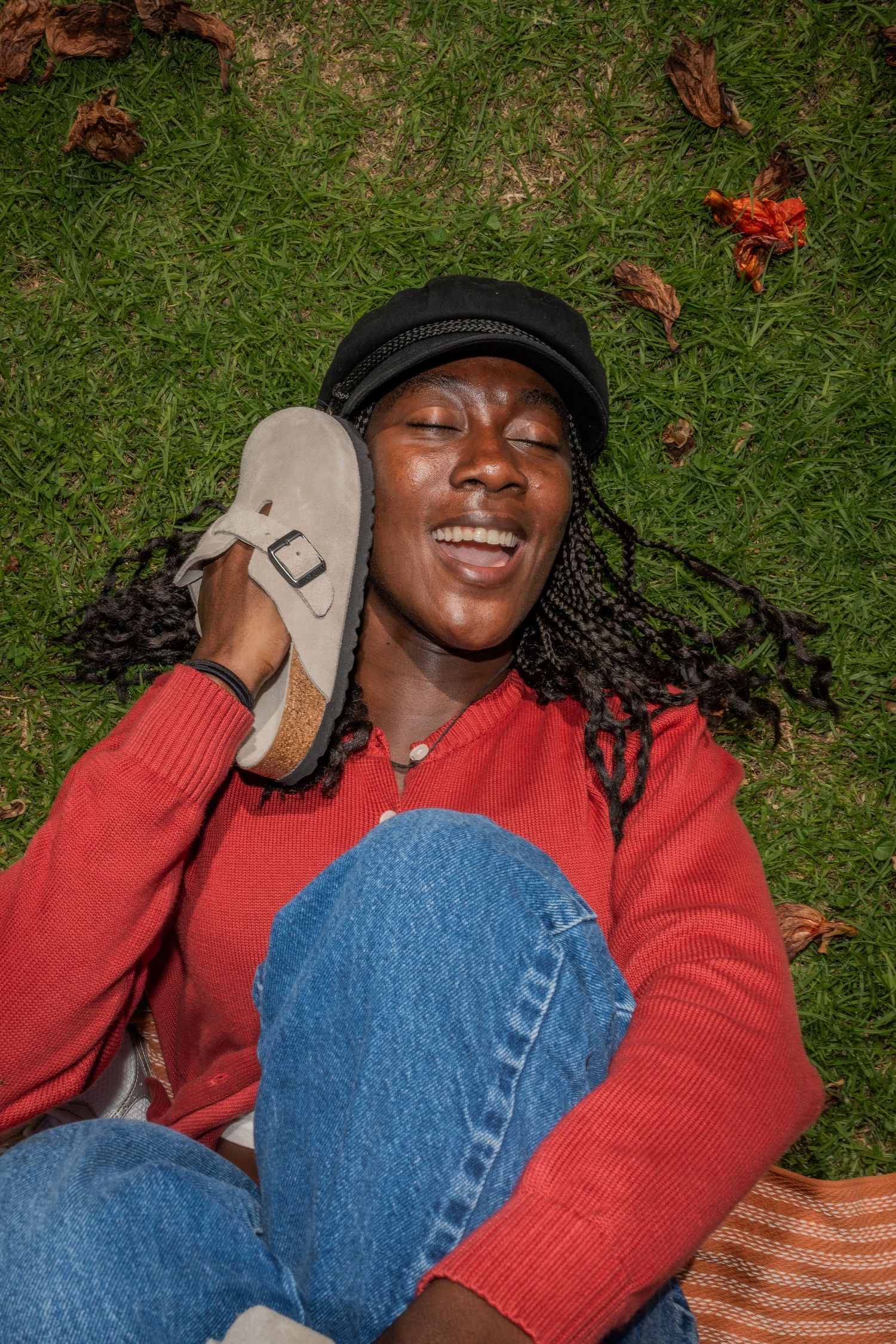 A woman with beautiful dark skin holds a tan Birkenstock sandal up to her face like a telephone with a big smile