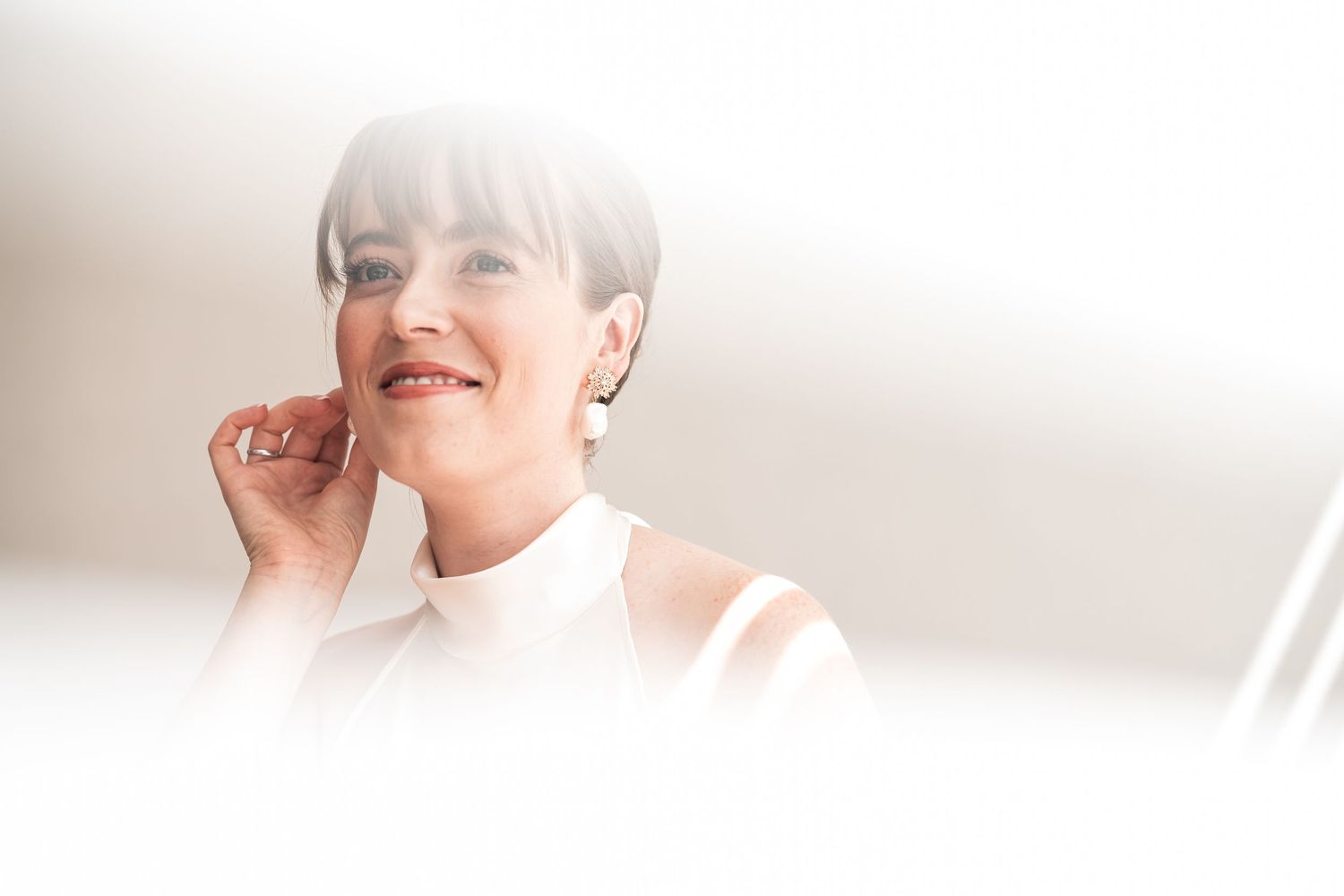 A close-up portrait in soft lighting shows a person in white wearing a minimalist hairstyle.