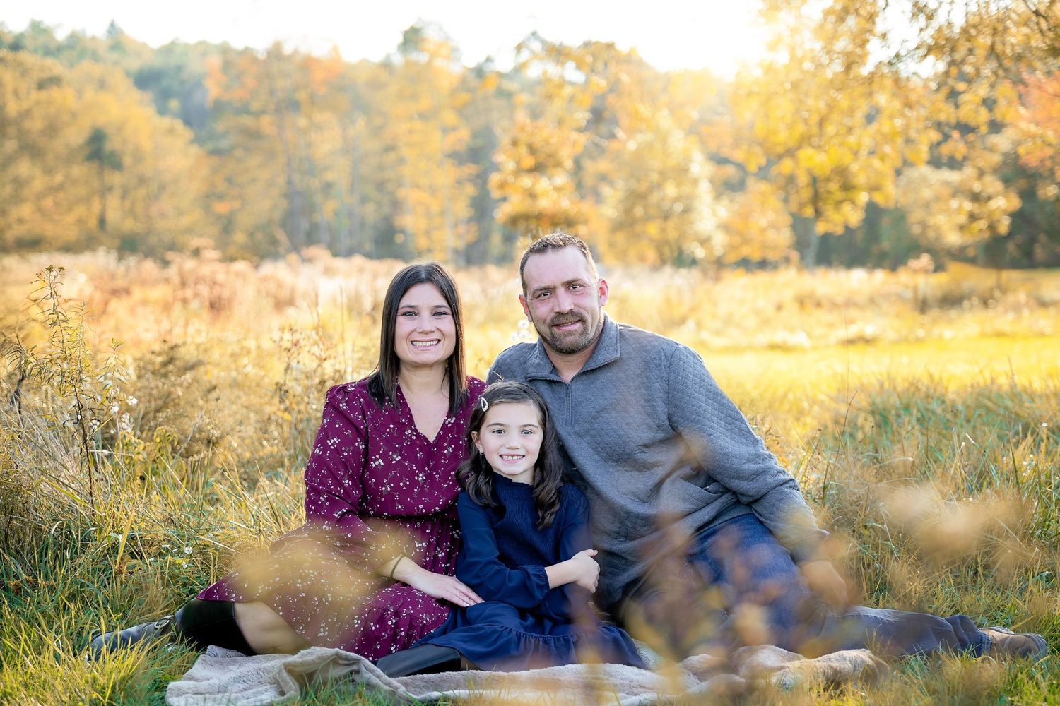 Family of three poses for outdoor portrait during golden sunset in field.