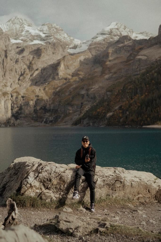 Person sitting on rock overlooking turquoise lake with snow-capped mountains in background.