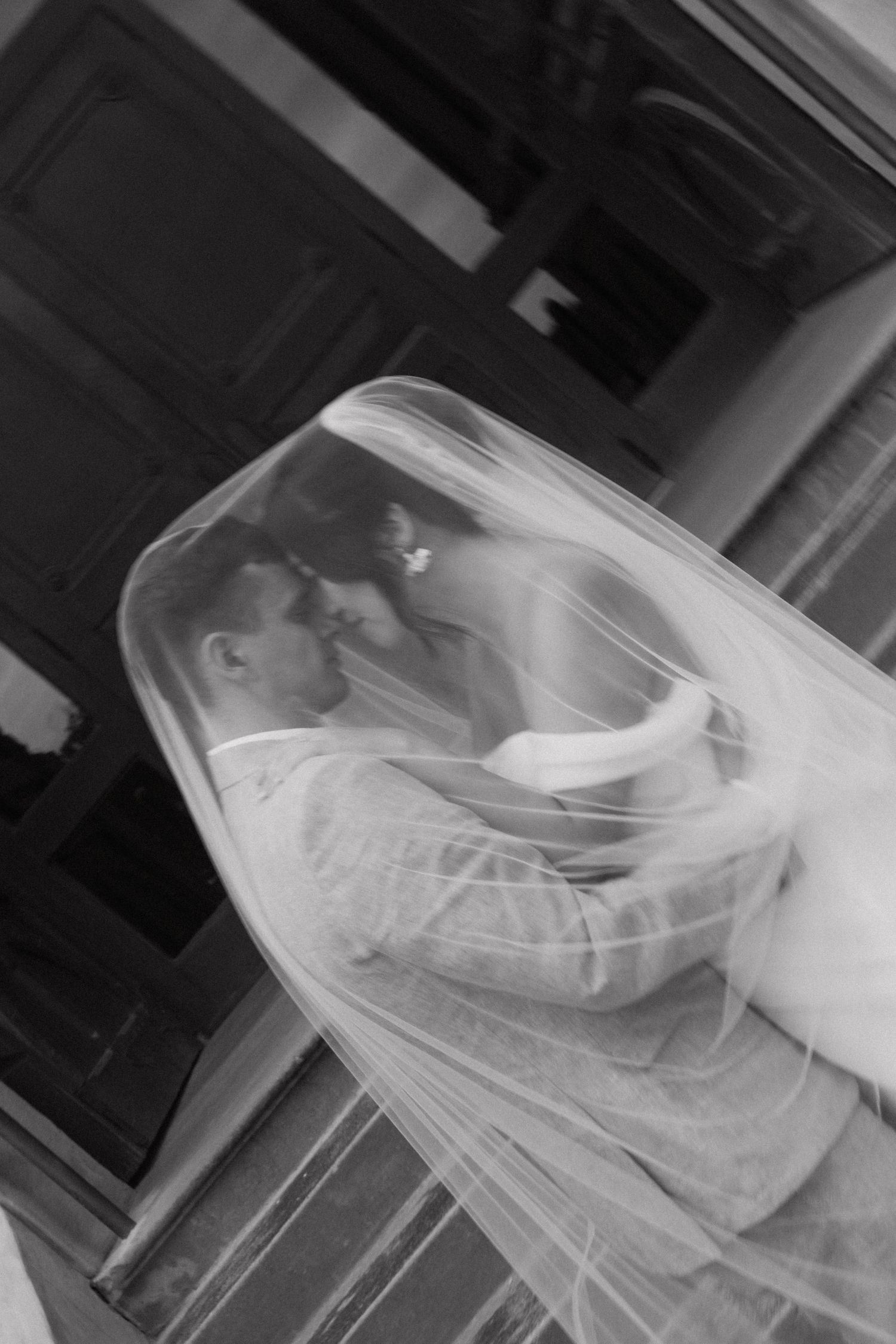 Black and white photo of bride and groom sharing a kiss under a flowing wedding veil.