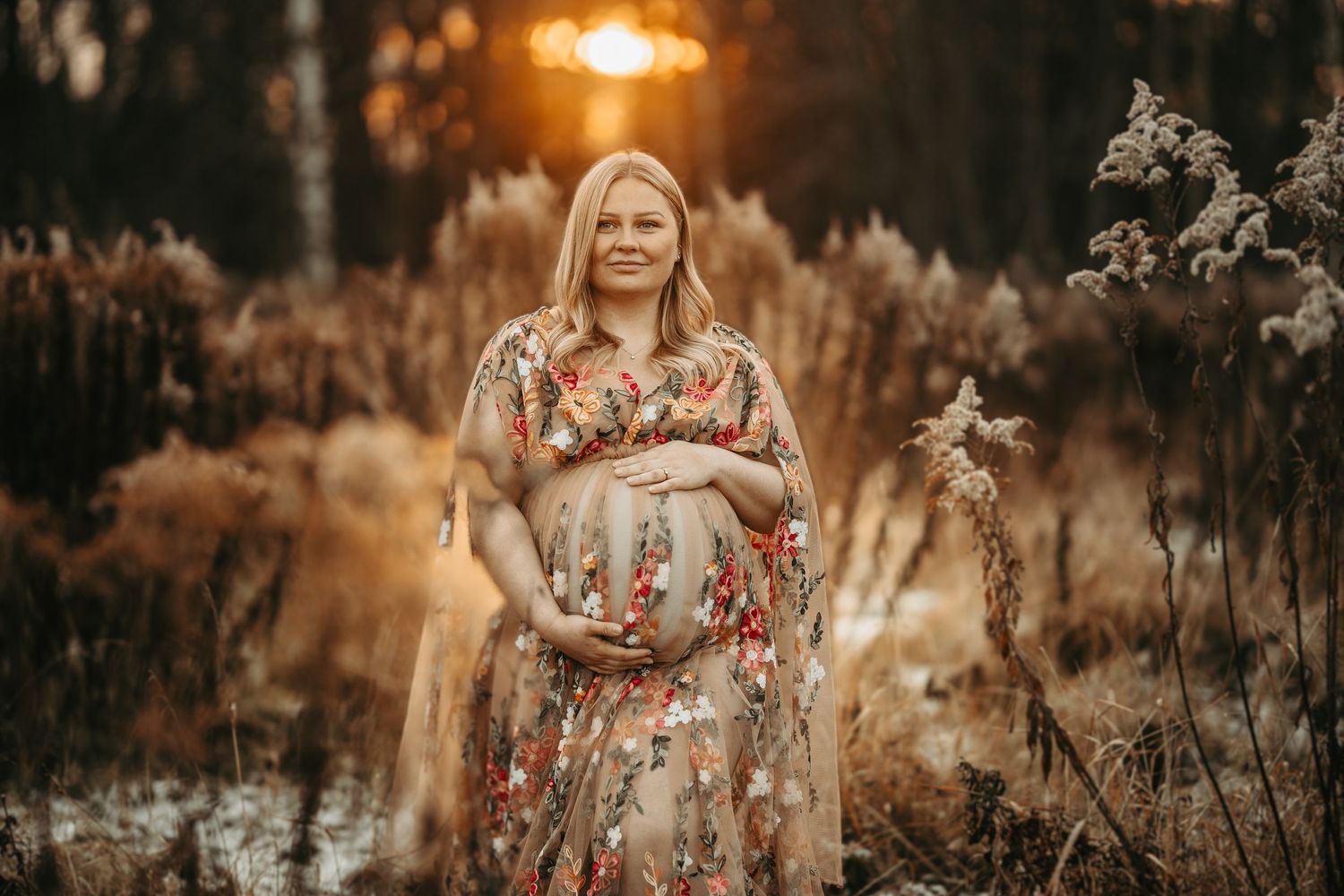 Ethereal maternity photo in a floral dress at golden hour in autumn field with tall grasses.