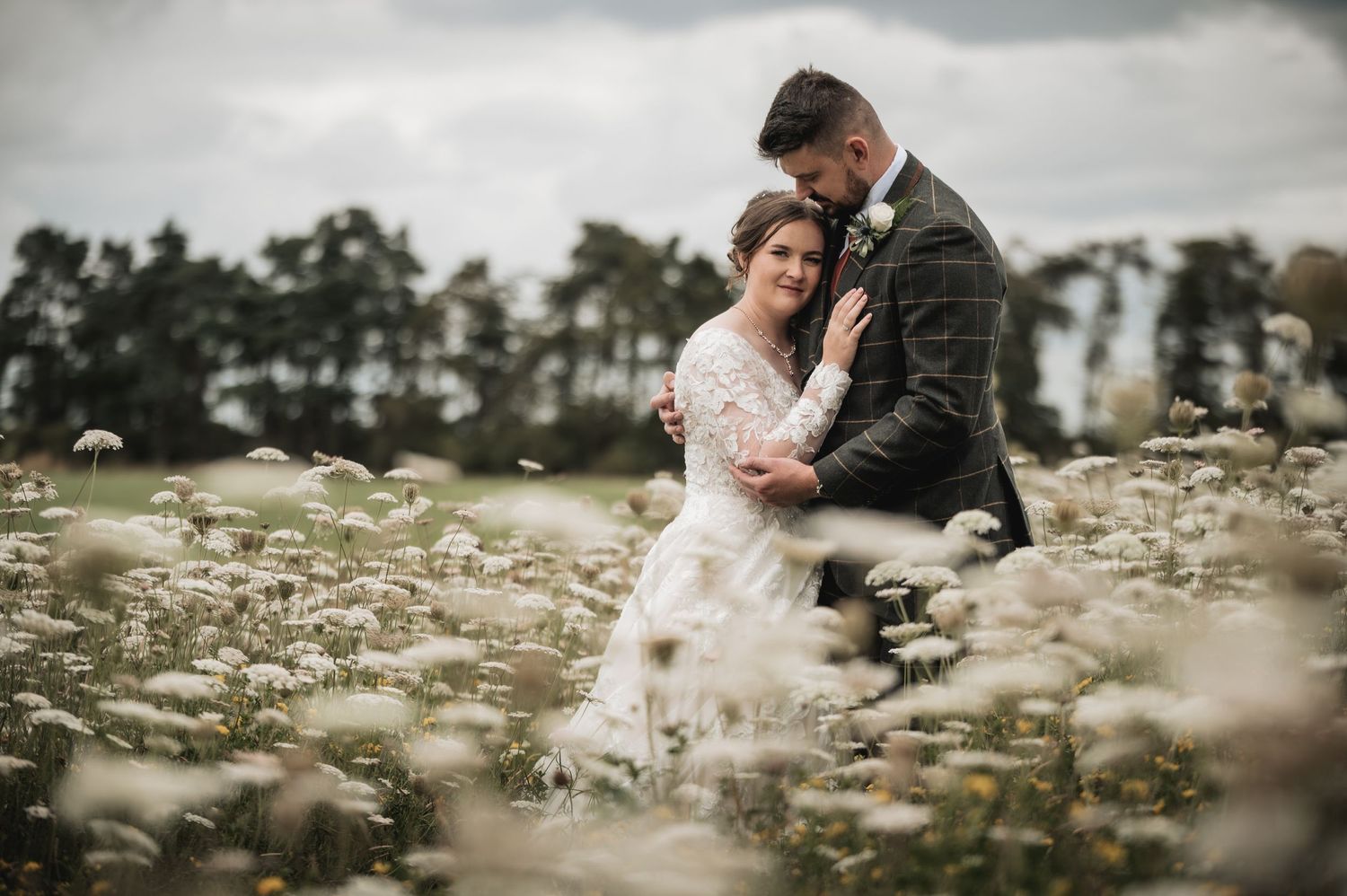 A romantic wedding photograph showing a couple embracing in a field of white wildflowers under a moody sky at Runningburn, Kelso