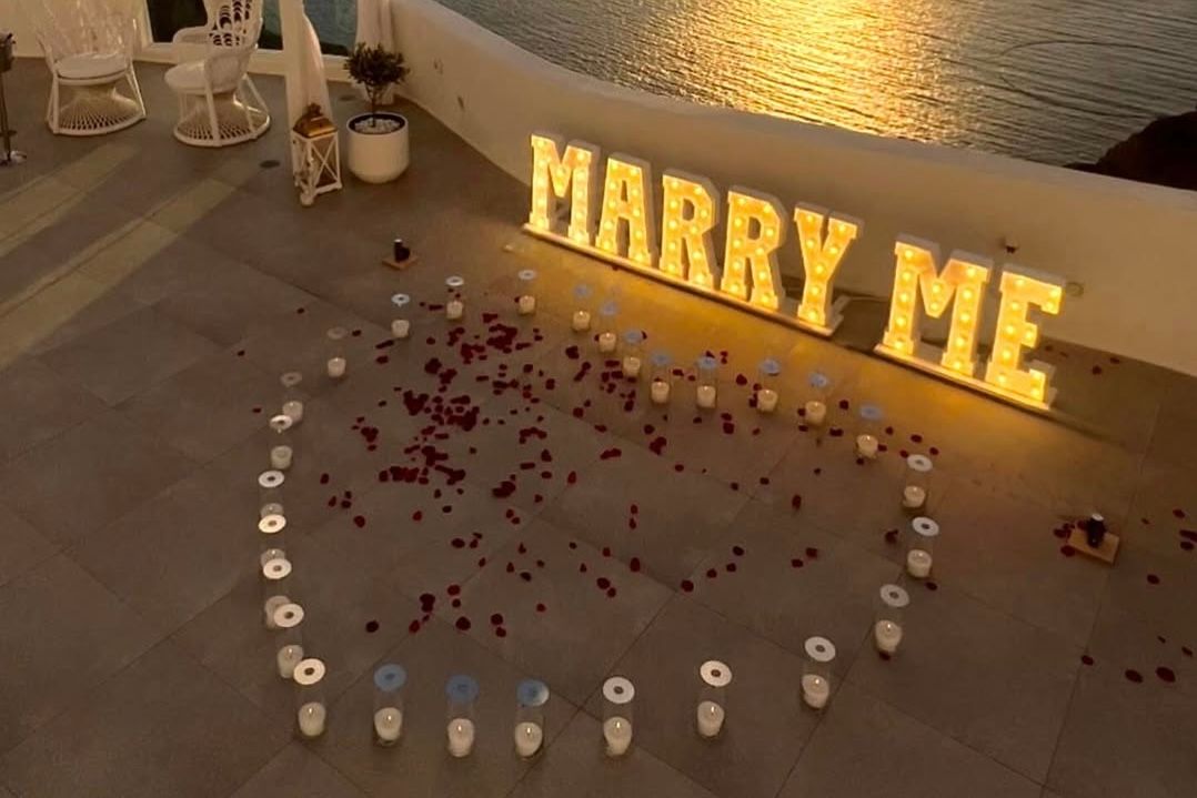 Glowing 'Marry Me' sign with candle heart and rose petals at sunset.