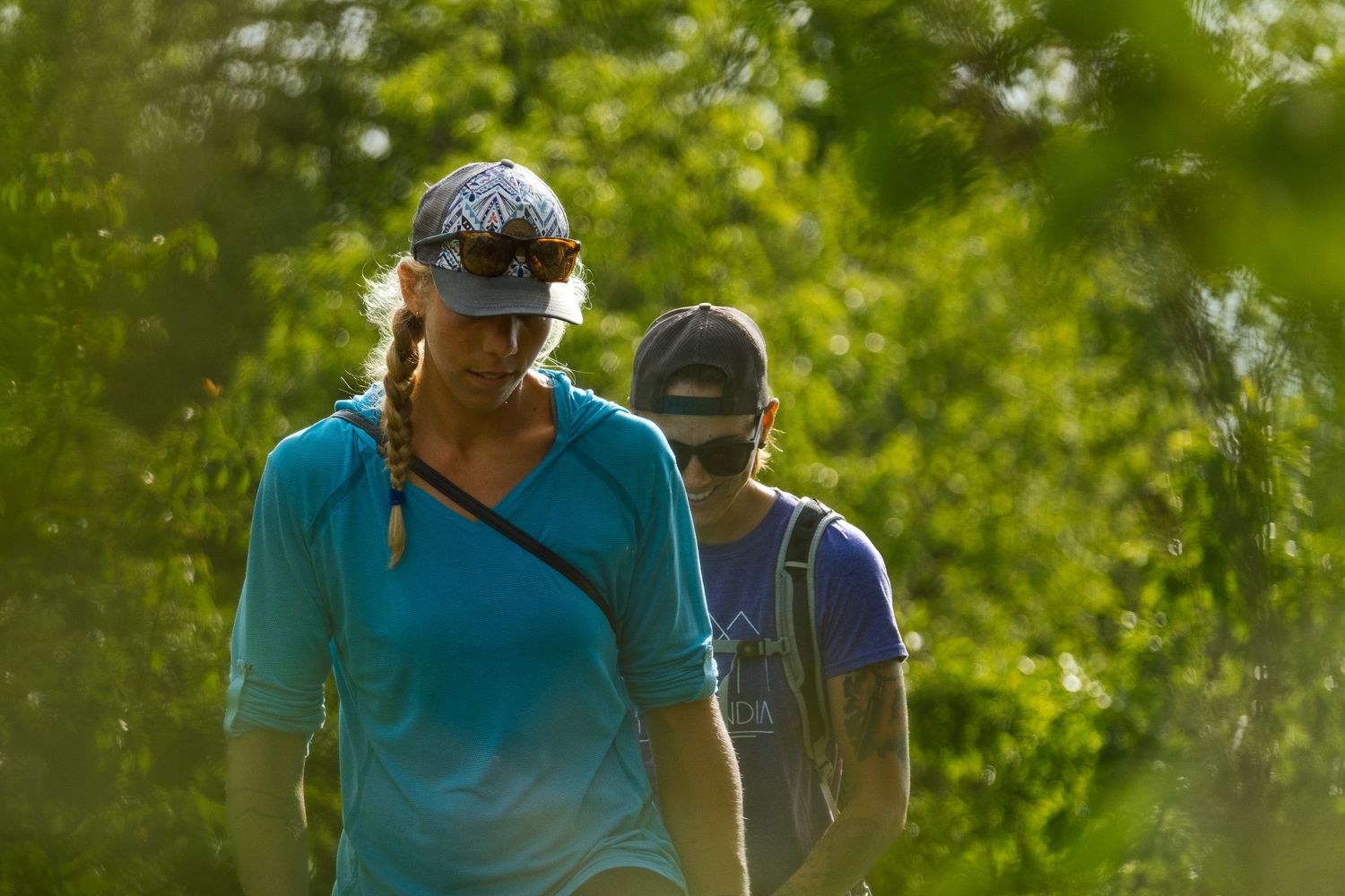 Two hikers walk along a forest trail on a sunny day wearing casual outdoor clothing and hats.
