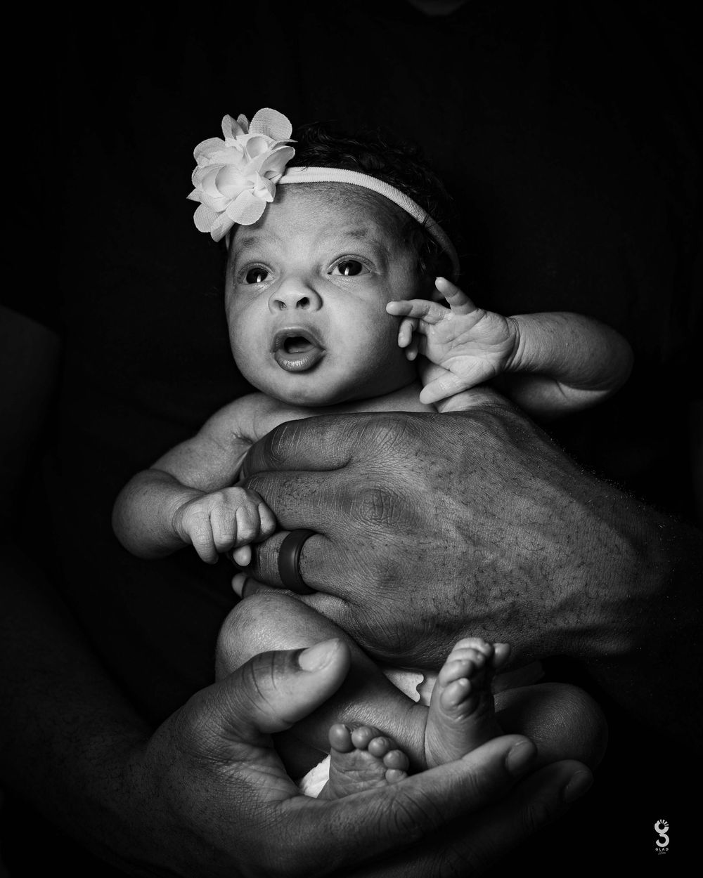 Sweet newborn baby wearing white floral headband lies cradled in adult hands in dramatic black and white portrait.