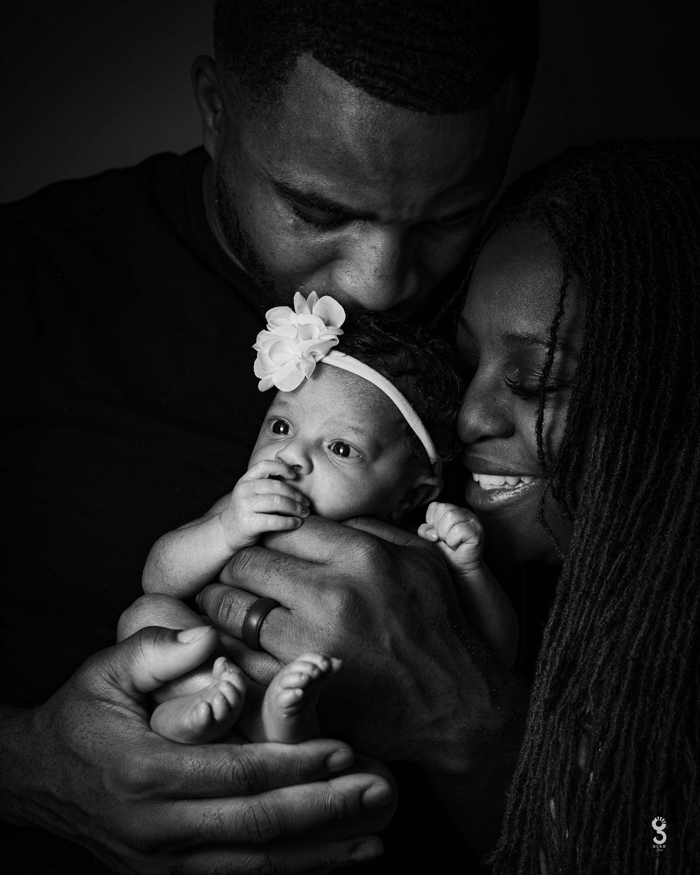 Black and white portrait of parents holding their small baby with a white flower headband in an intimate family moment.
