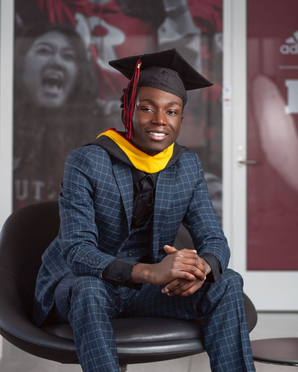 Graduate in navy plaid suit and gold stole sits in dark chair wearing academic cap and gown for graduation portrait.
