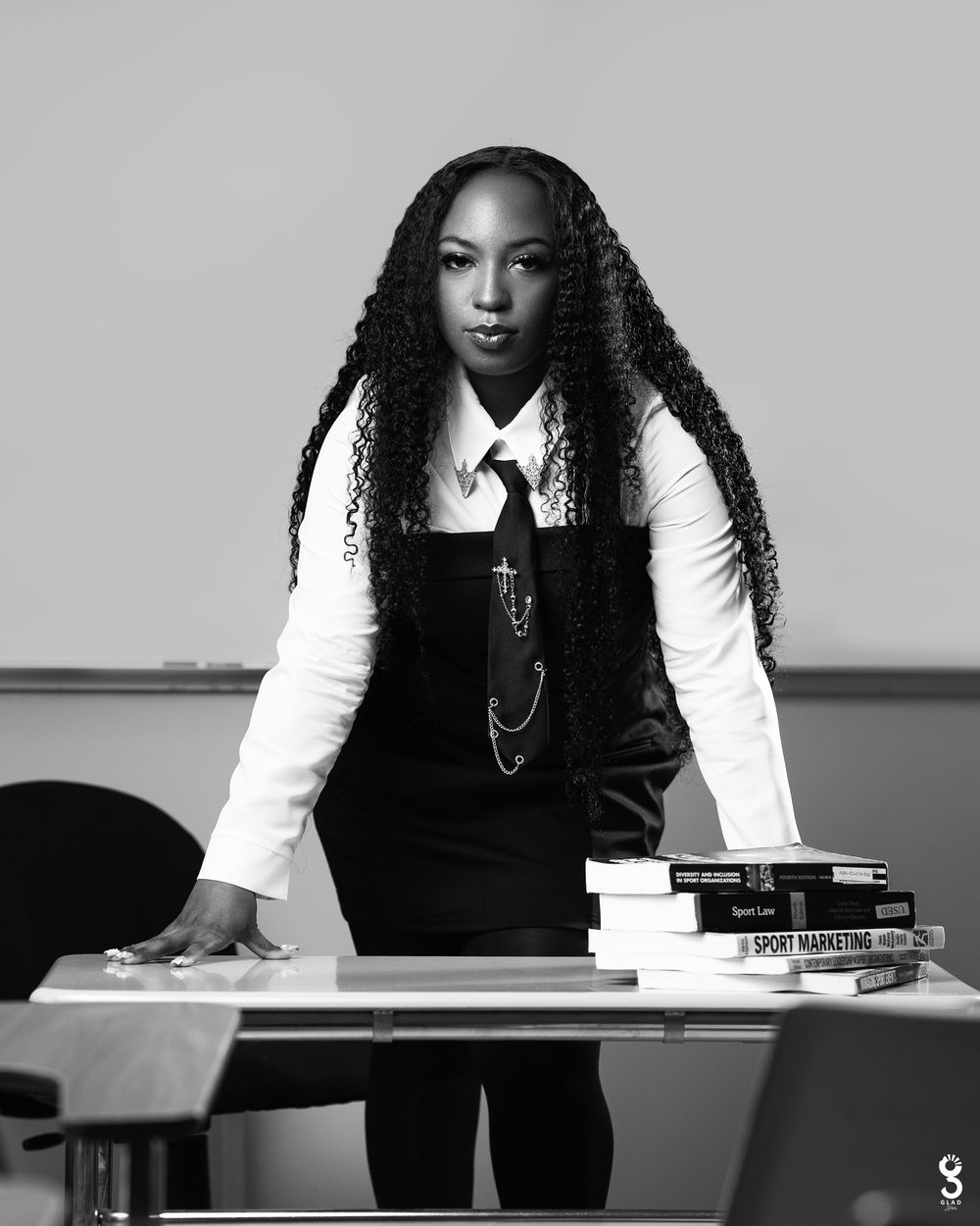 Monochrome studio portrait of a professional in a white shirt and black overalls leaning on a desk with stacked books.