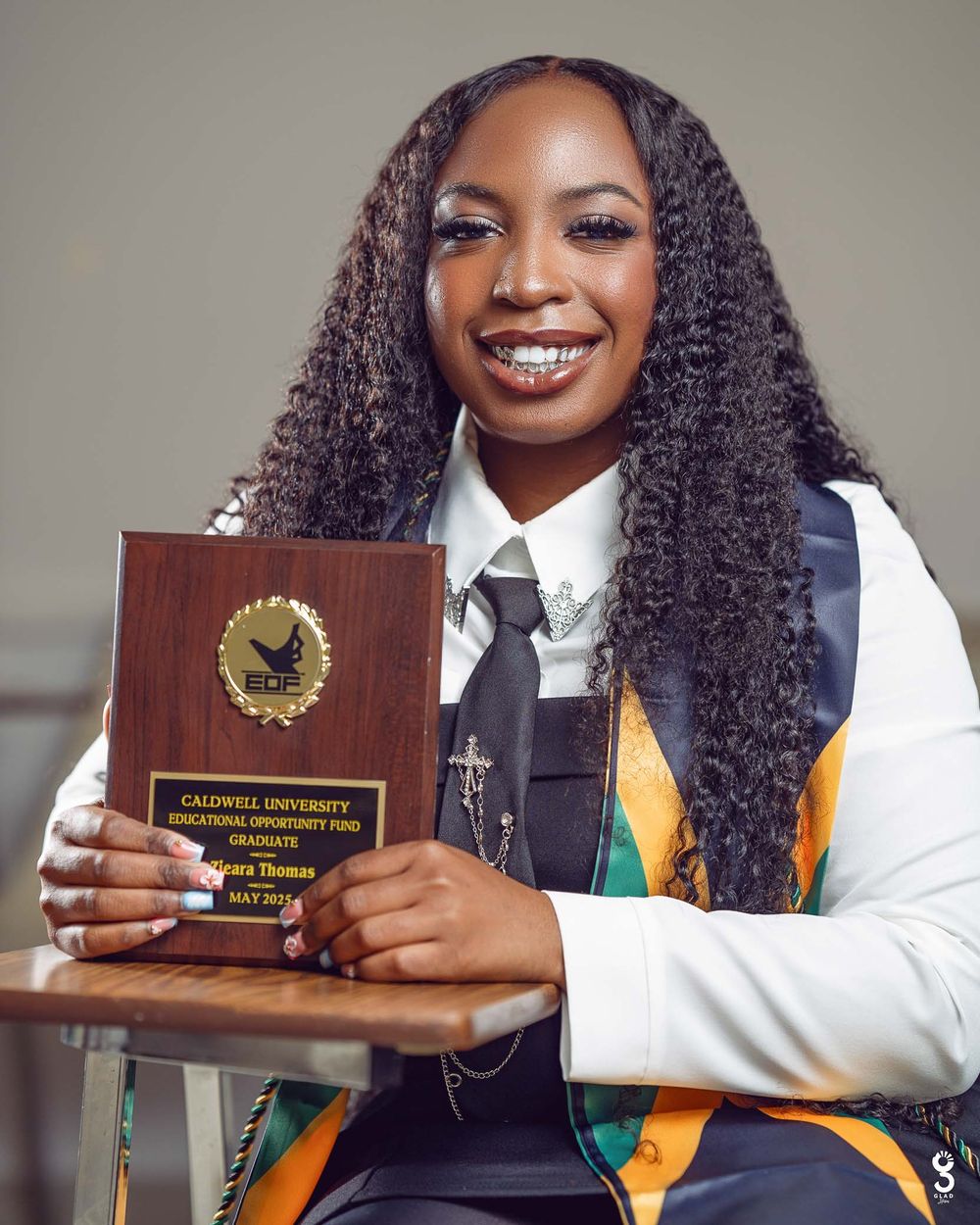 Graduate student holding a wooden academic achievement plaque while wearing graduation stole and white shirt with tie.