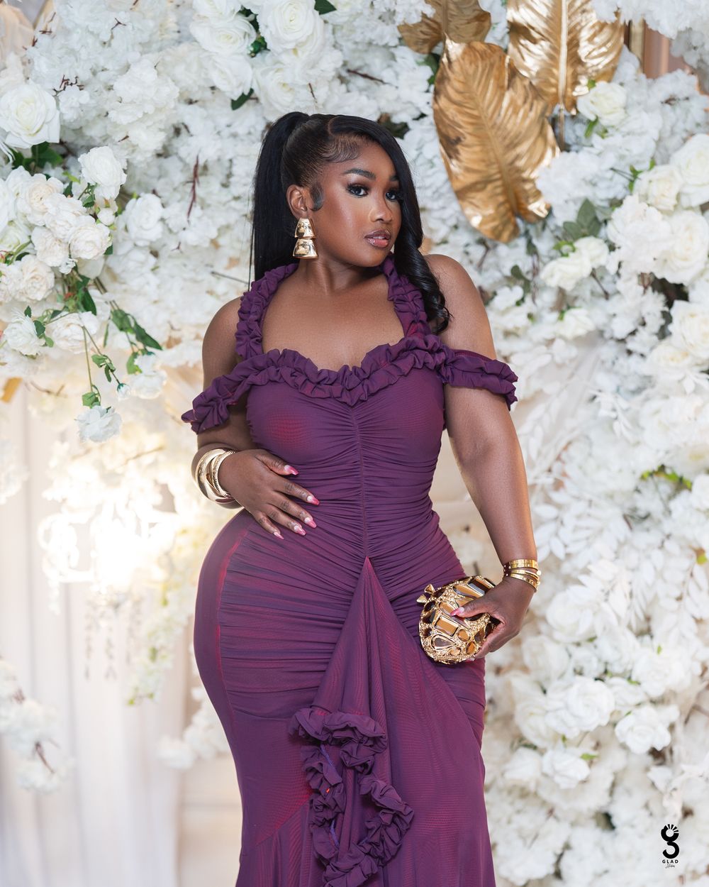 A stunning purple ruched dress with ruffled details is showcased against a backdrop of white floral decorations.