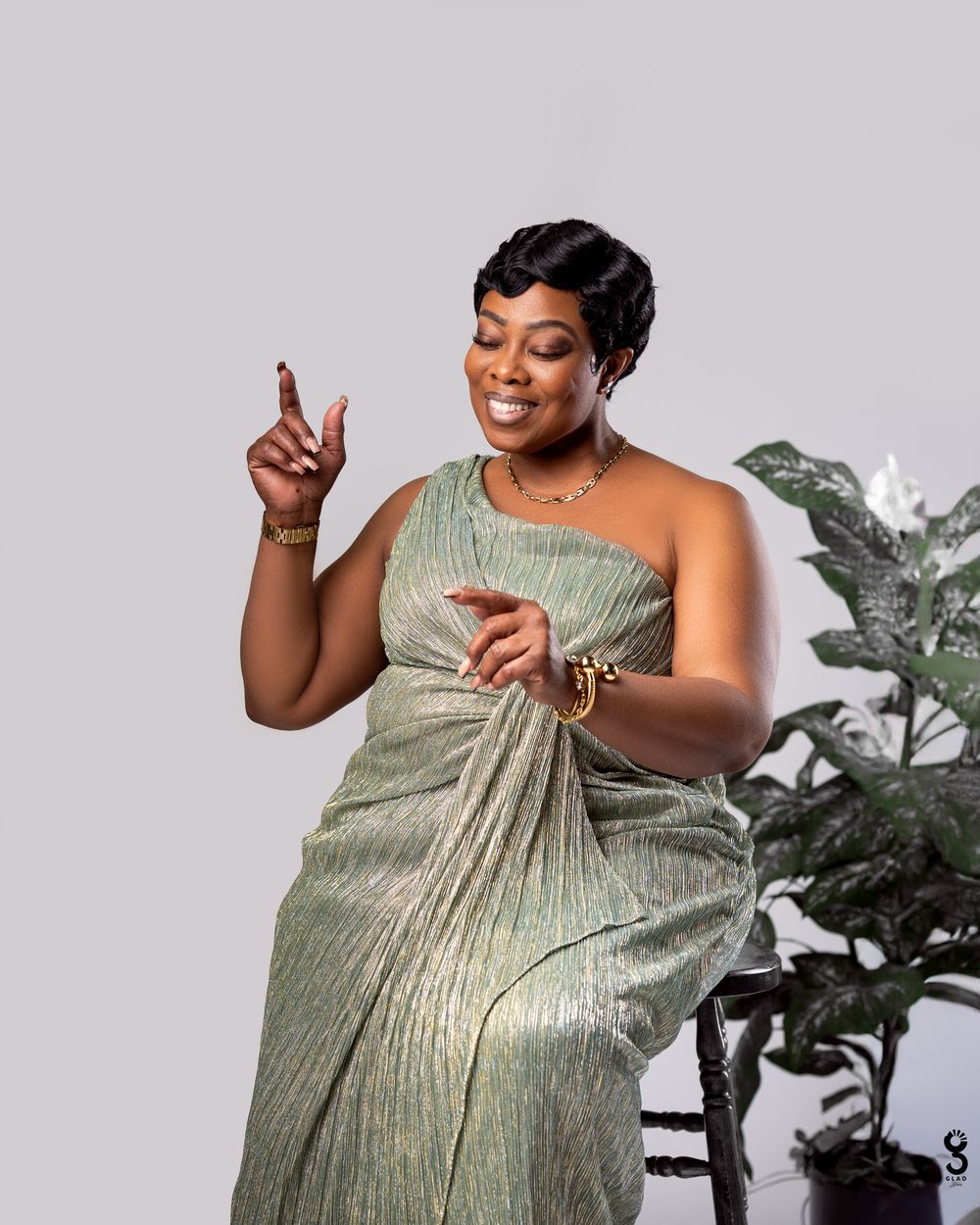 A person in a shimmering sage green pleated dress poses confidently against a light background with decorative plants.