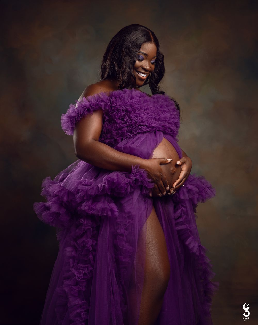 A glamorous maternity photoshoot featuring a flowing purple tulle gown against a dark background.
